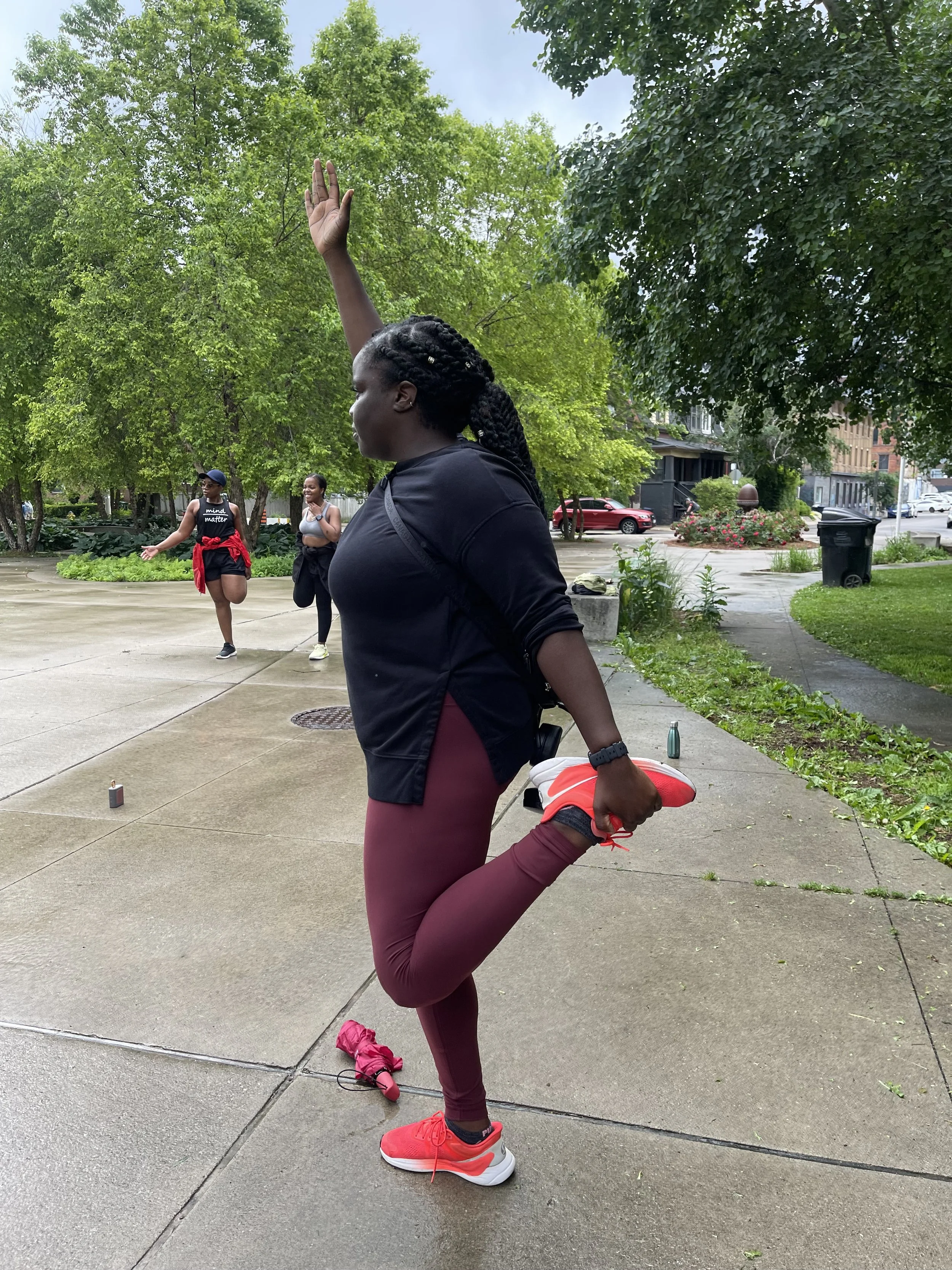 A woman in athletic wear is stretching her leg while standing on a concrete sidewalk in a park or urban area with trees and a few people in the background.