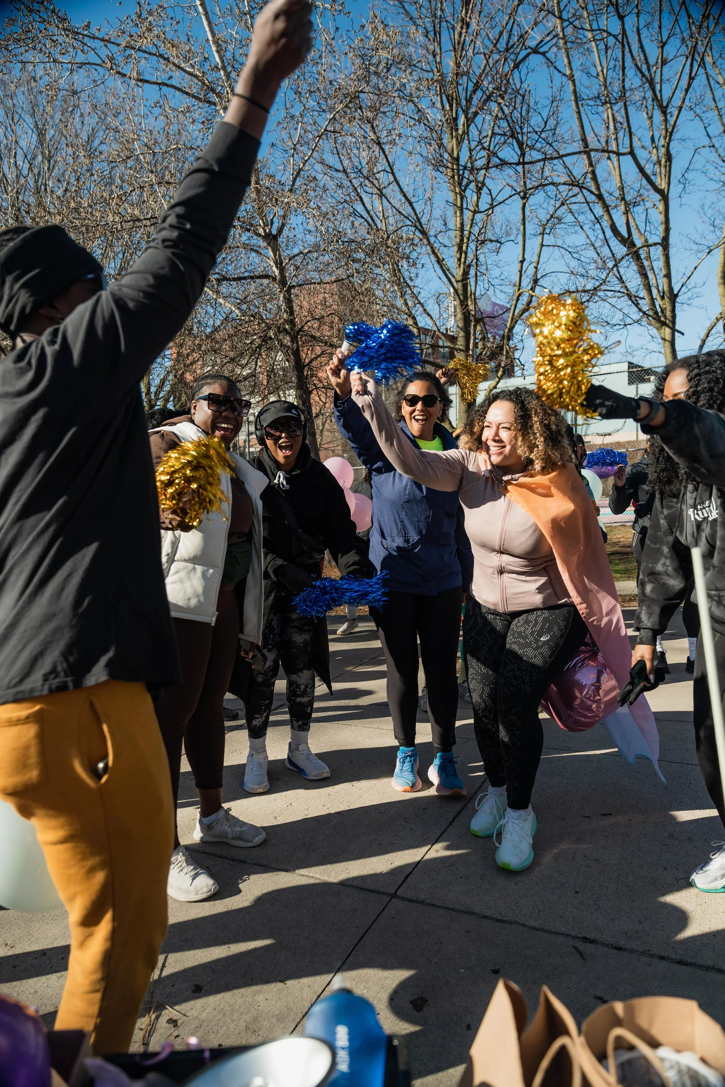 Group of women outdoors celebrating with colorful pom-poms and balloons on a sunny day.