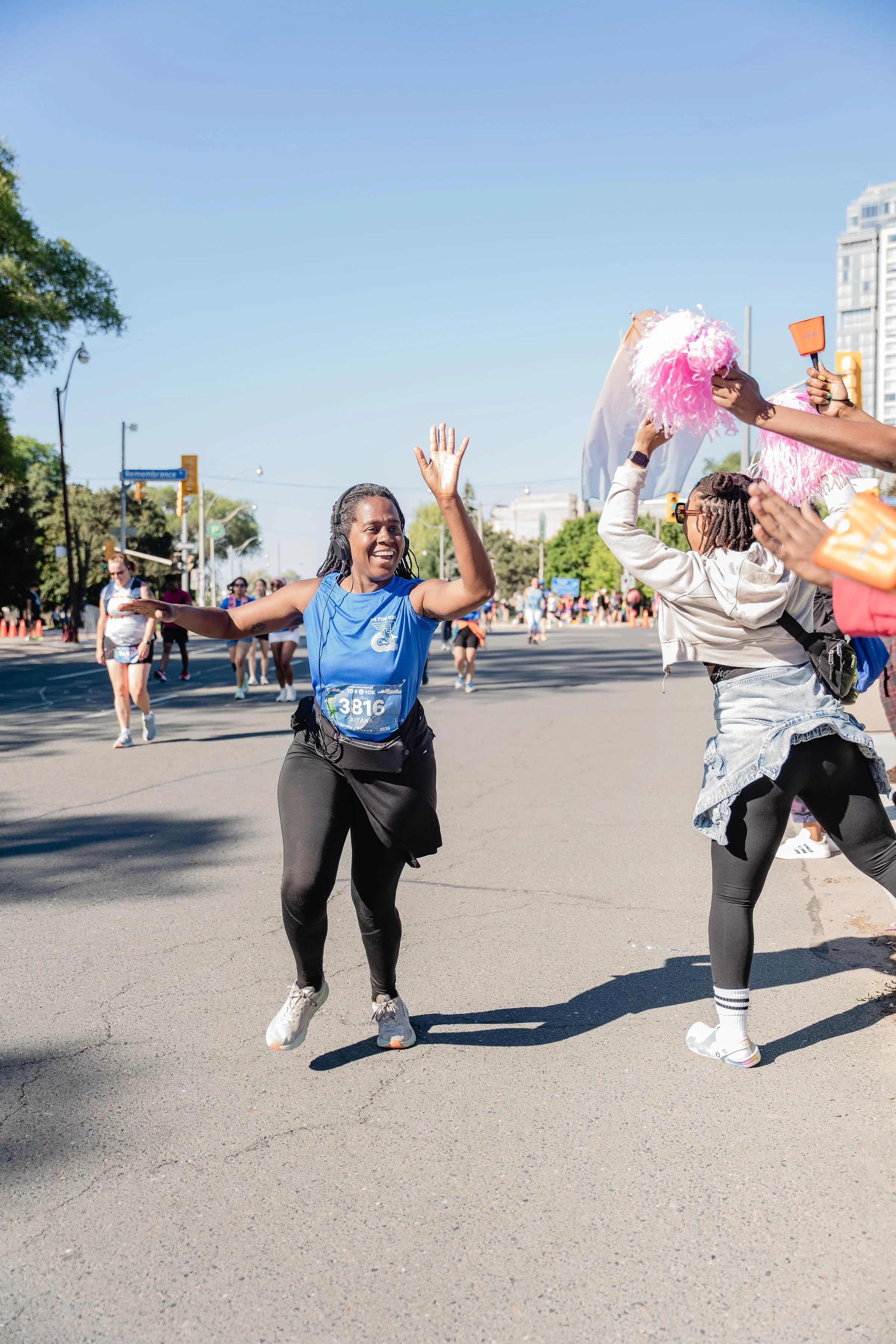A woman in a blue race shirt and black leggings high-fives a helper with pink pom-poms during a marathon or fun run event on a city street.