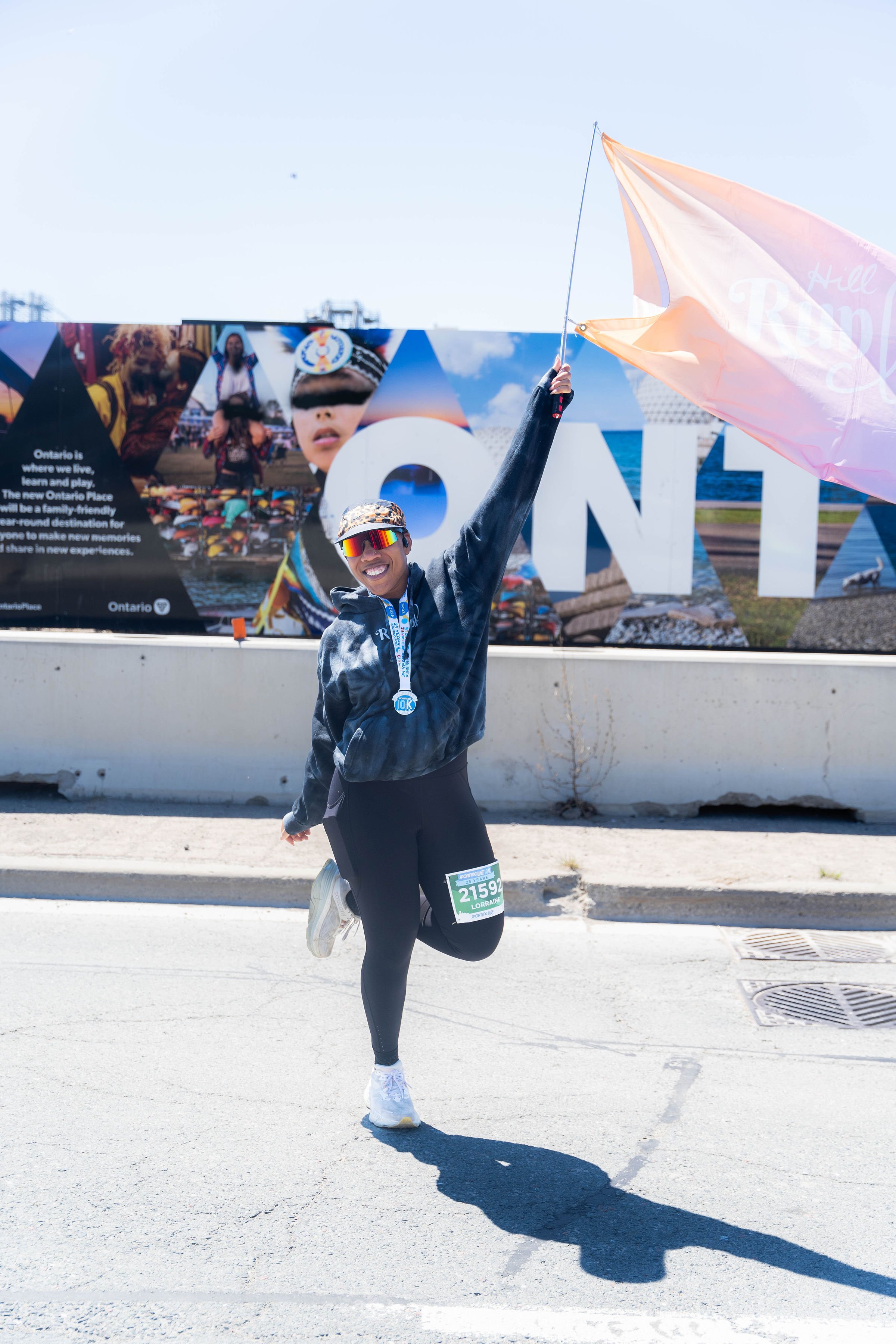 A woman celebrating at a marathon, holding a pink flag in one hand and wearing a medal, running on a city street with a large billboard in the background.