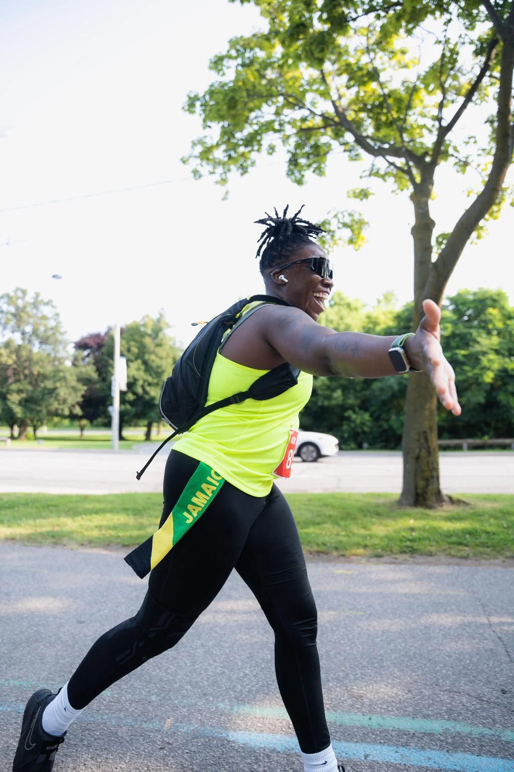 A smiling woman in athletic gear extending her hand for a high five during a race, with trees and a street in the background.
