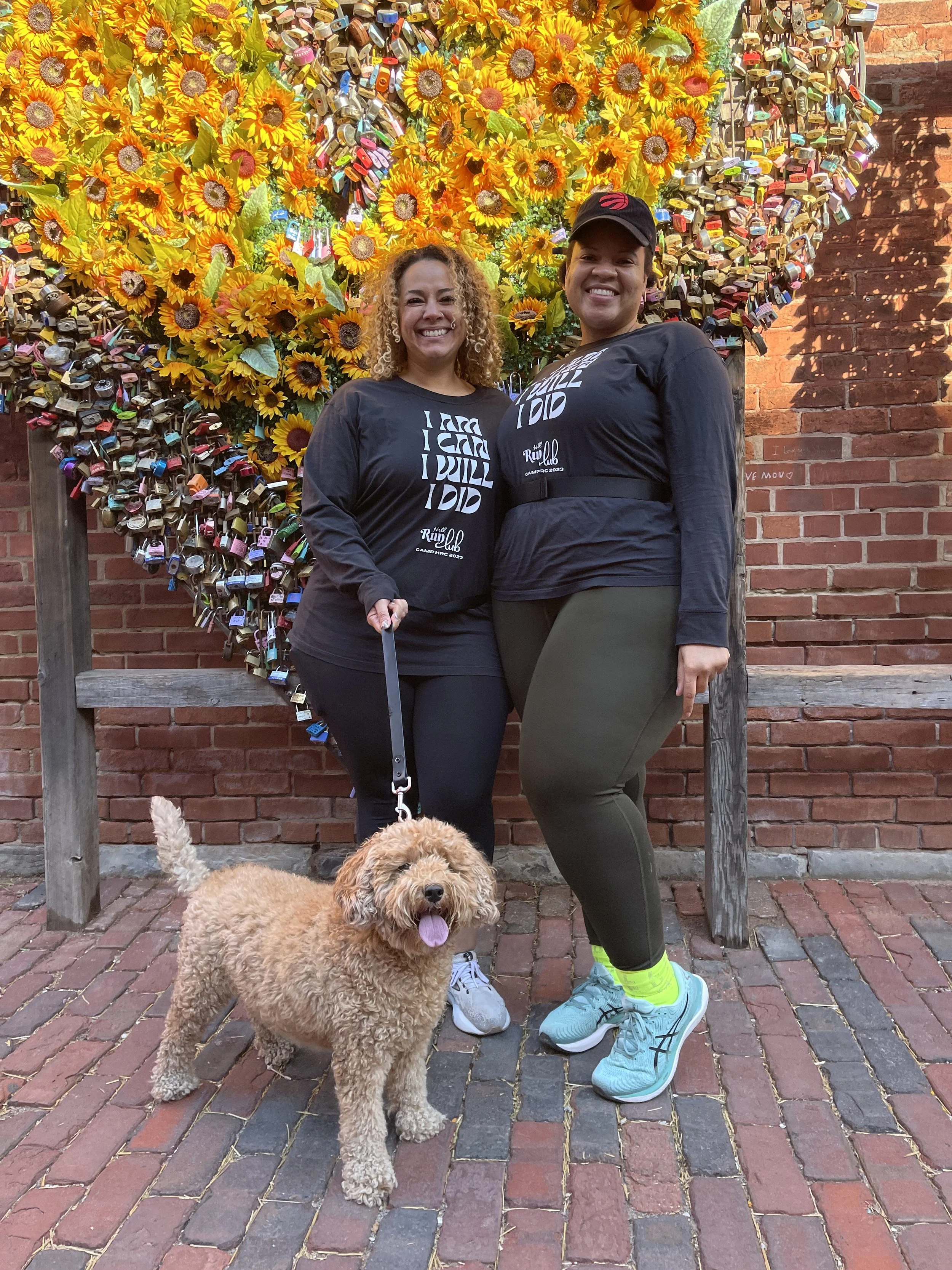 Two women smiling and standing on a brick walkway in front of a wall decorated with thousands of padlocks and yellow sunflowers. One woman is holding a leash attached to a curly-haired dog. Both women are wearing matching black long-sleeve shirts.