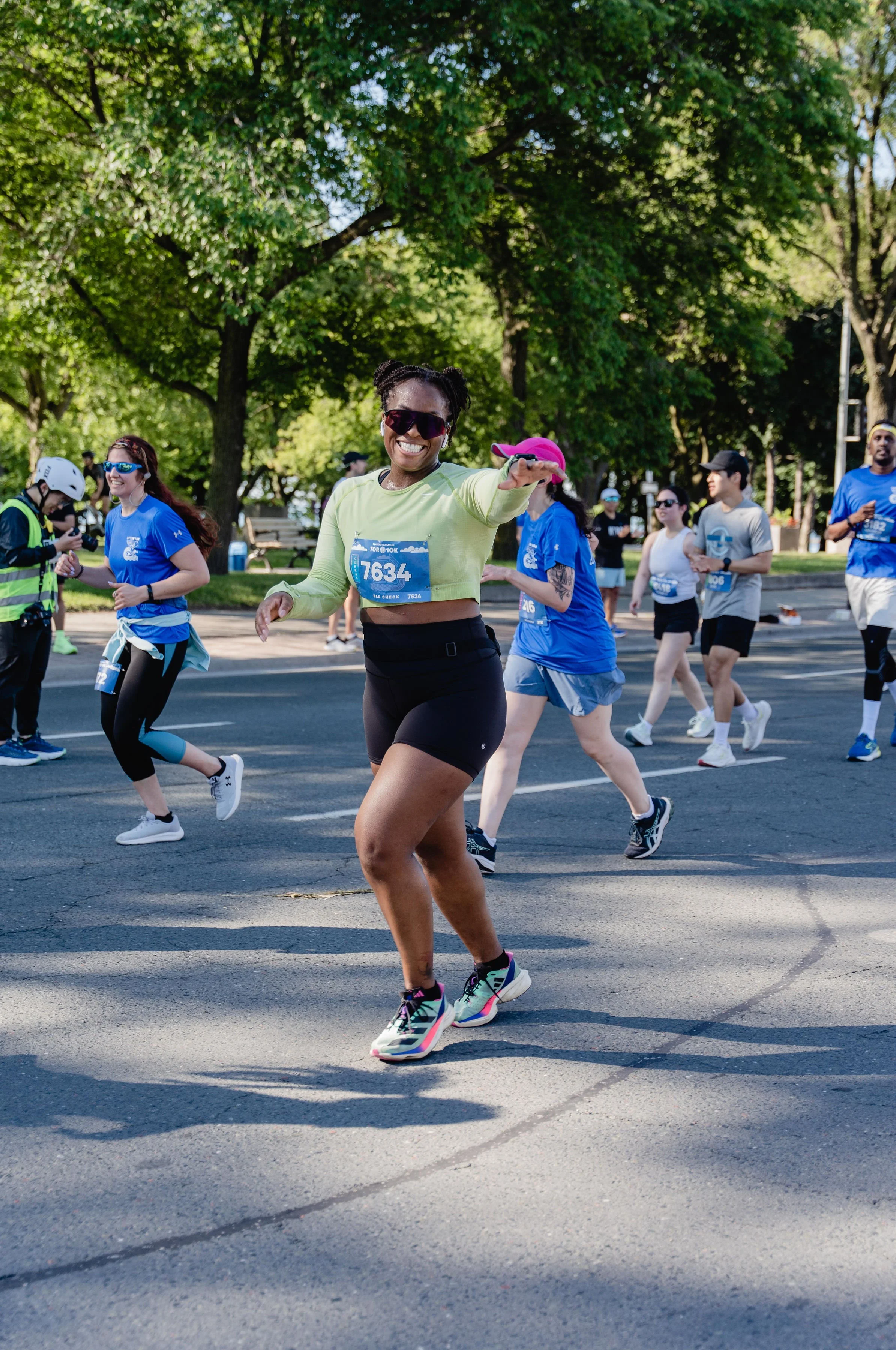 Smiling woman in lime green shirt and black shorts running in a marathon, surrounded by other runners in blue and gray shirts, on a tree-lined street.