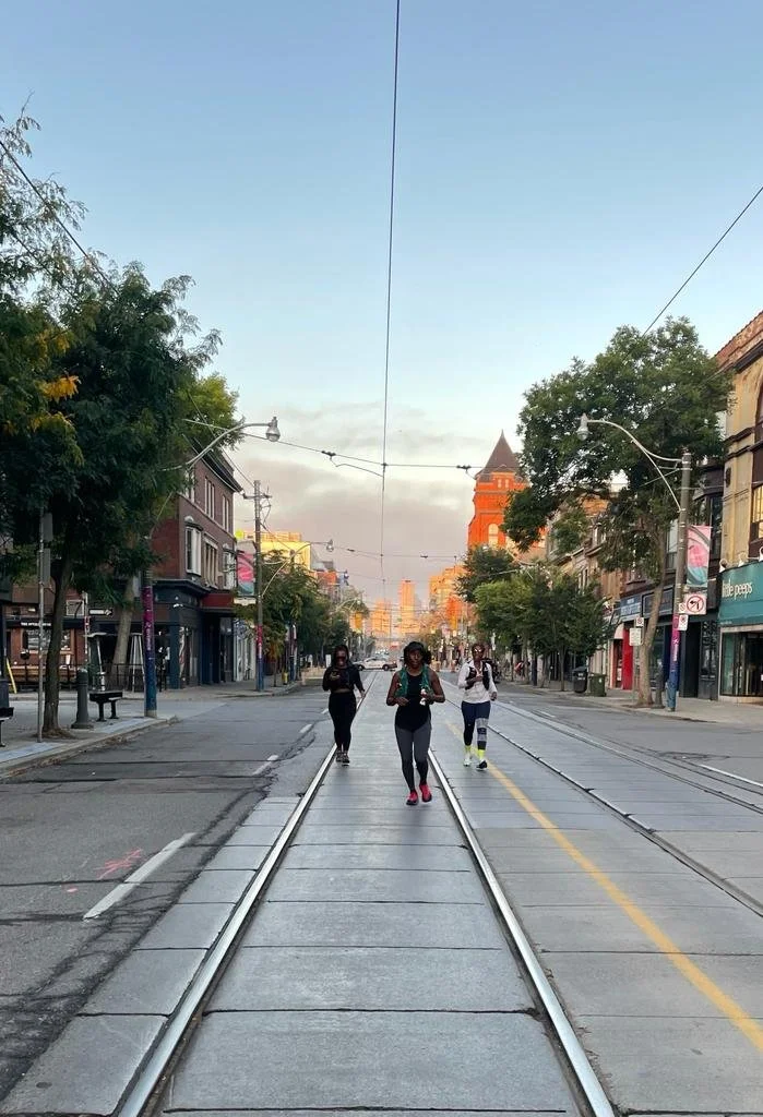 Three Black women running on a city street in Toronto with streetcar tracks, city buildings, trees, and a tall tower in the background.