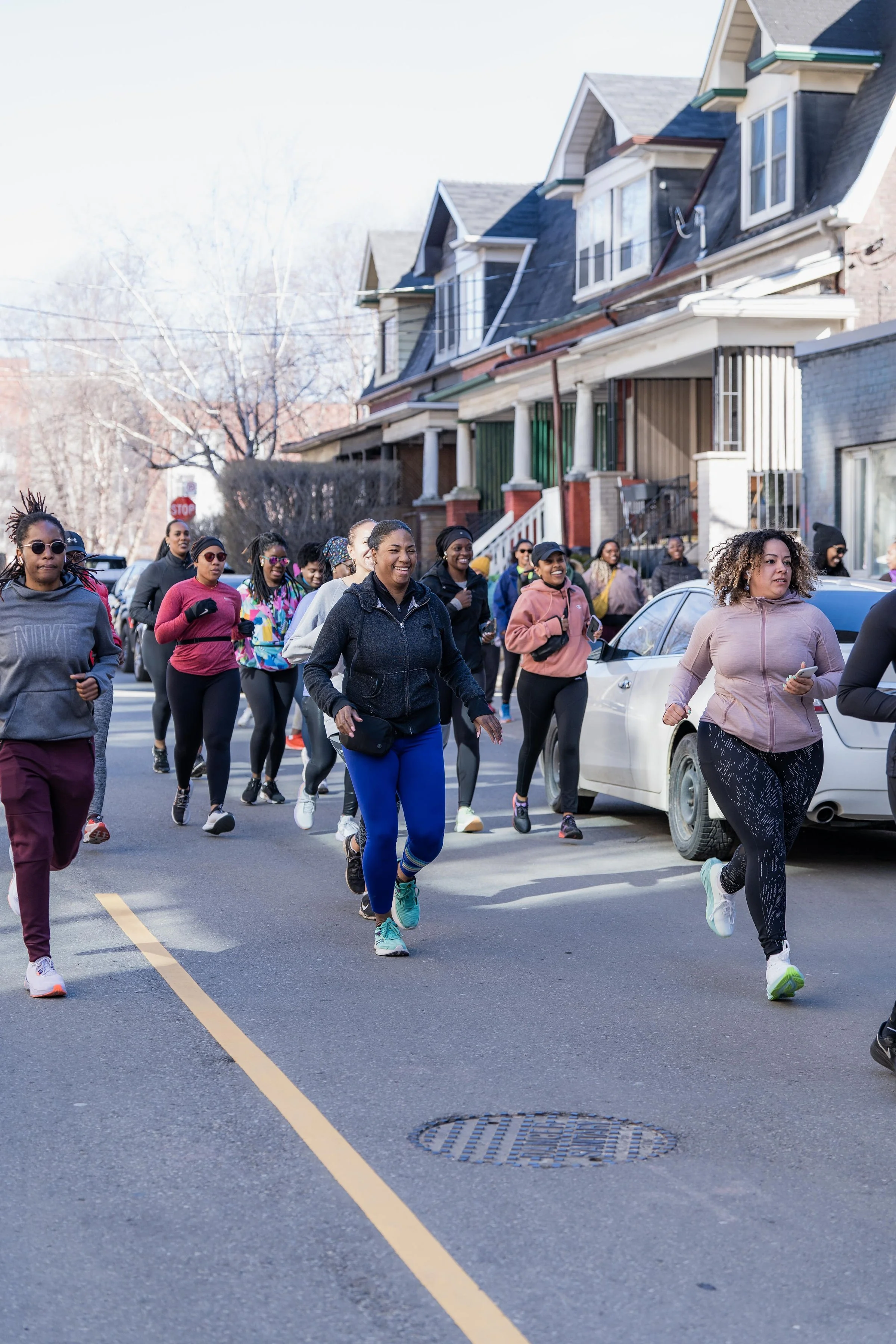 A group of women jogging or running together on a city street, wearing athletic clothing, with houses and cars visible in the background.
