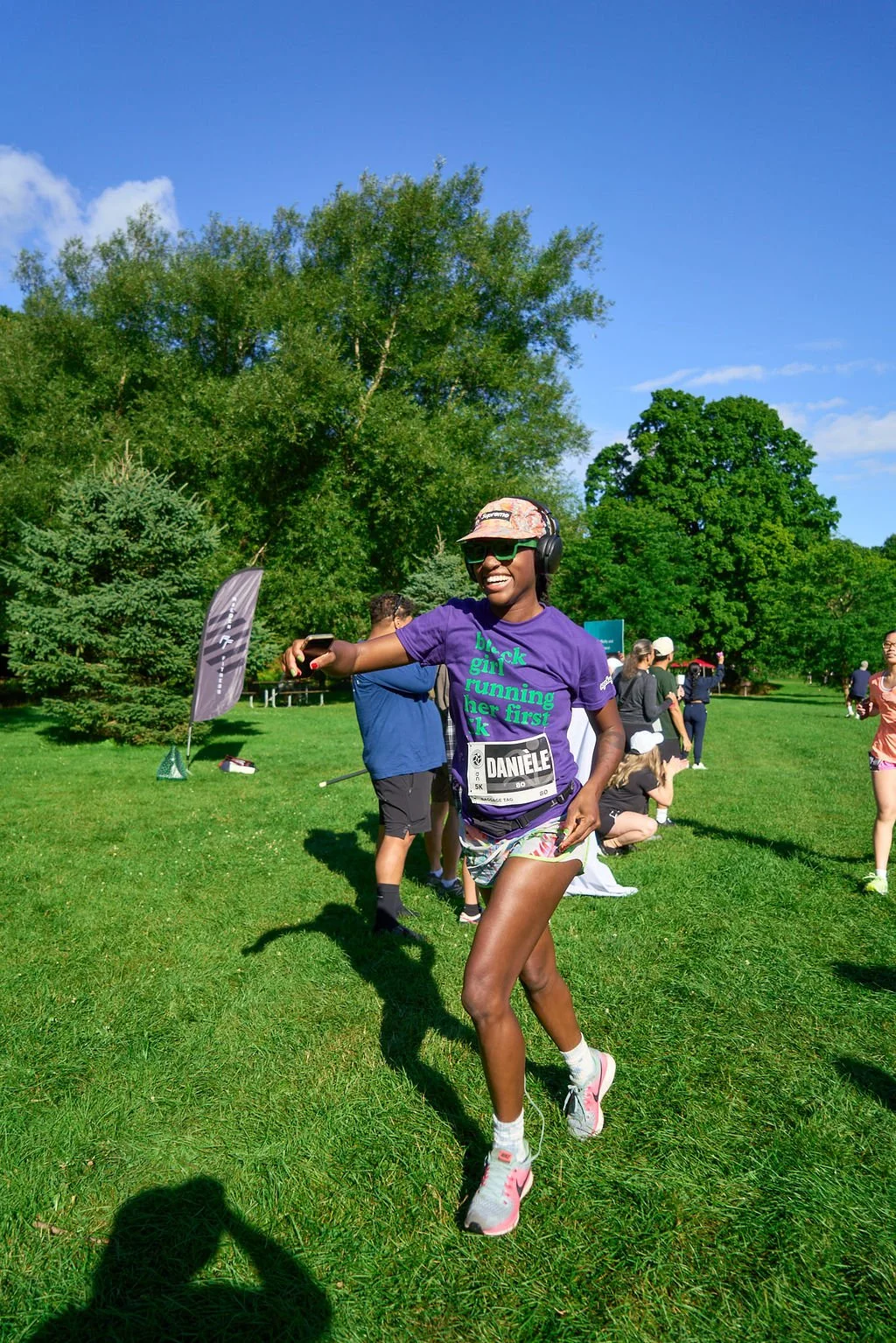 A woman running outdoors during a race, wearing a purple T-shirt, shorts, sunglasses, a cap, and headphones, smiling and holding a phone in her right hand. Other people and trees are visible in the background on a sunny day.