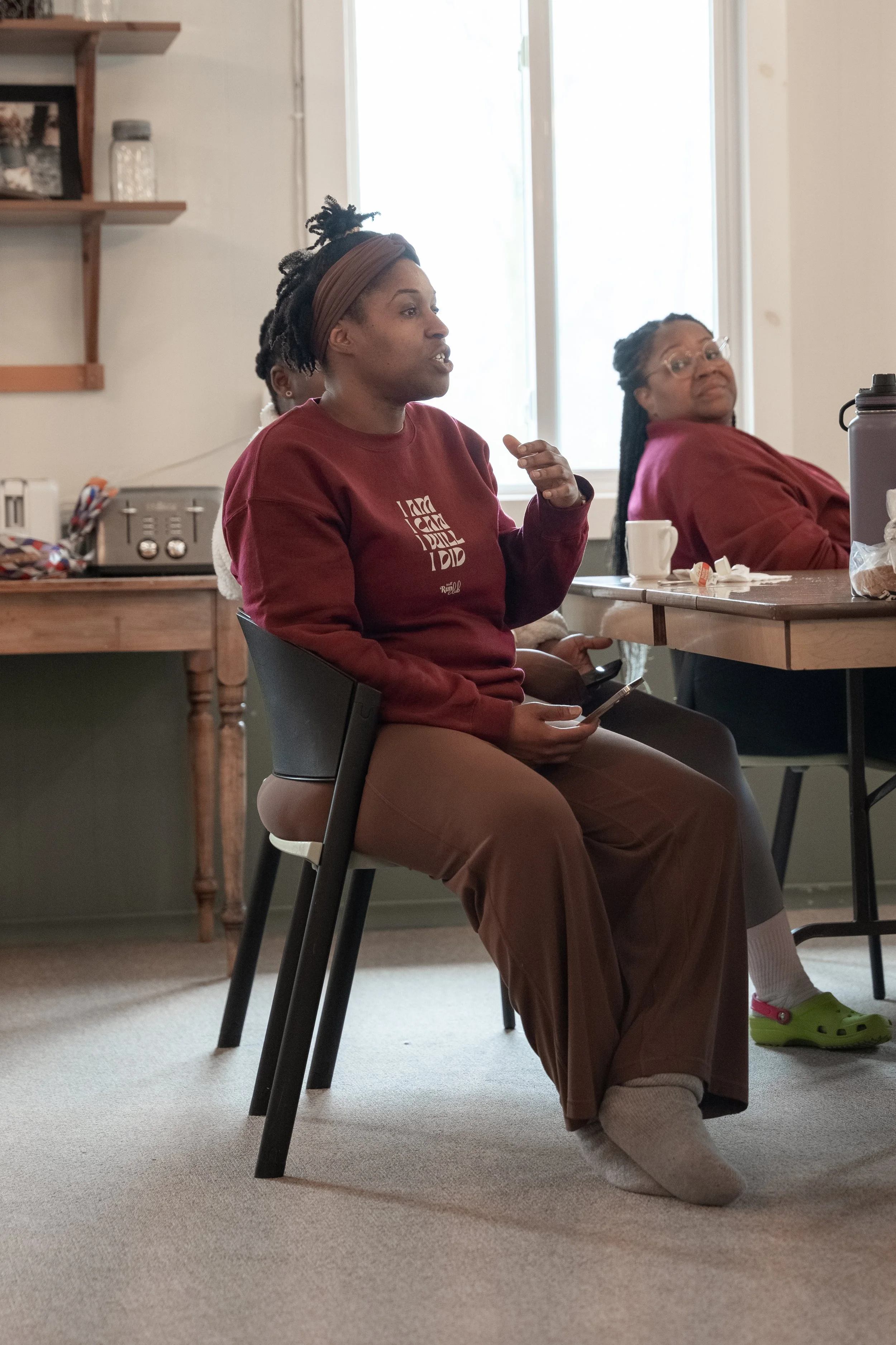 A woman with dreadlocks wearing a maroon sweatshirt and brown pants sitting on a chair, speaking and holding a phone, while two other women sit at a table in the background.