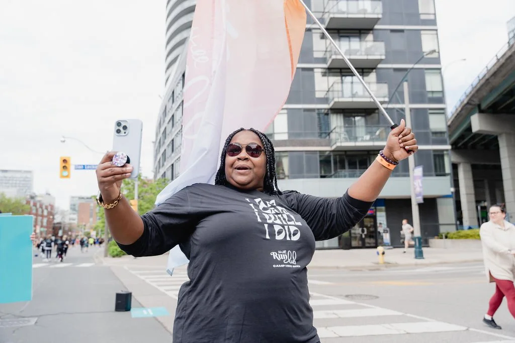 A woman smiling and taking a selfie with a smartphone on a city street, holding a flag, wearing sunglasses and a black t-shirt.
