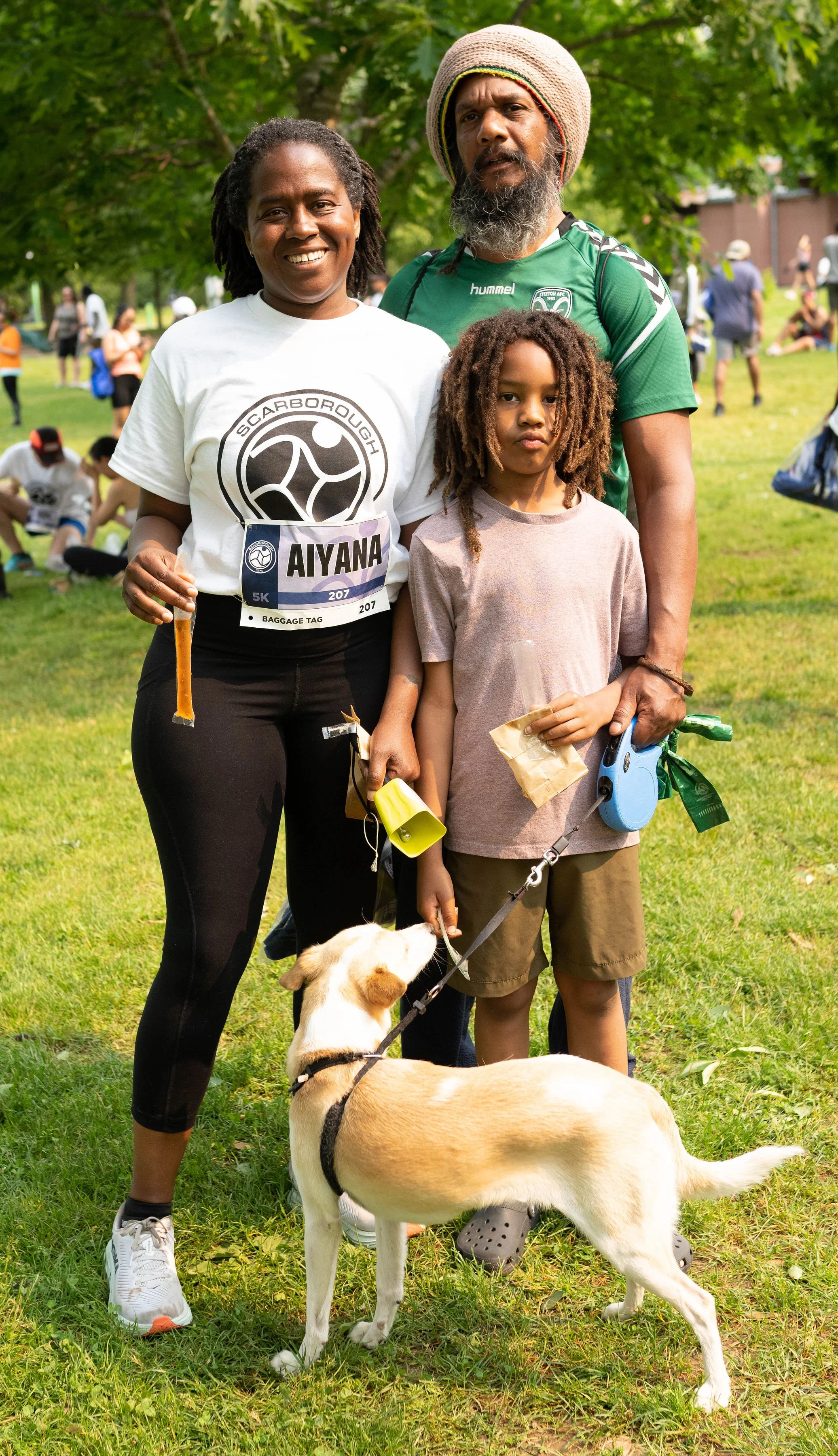 A family of three with a dog at an outdoor event, standing on grass with trees in the background, surrounded by other people. The woman holds a small stick, the man wears a hat, the child holds a paper bag, and the dog is on a leash.