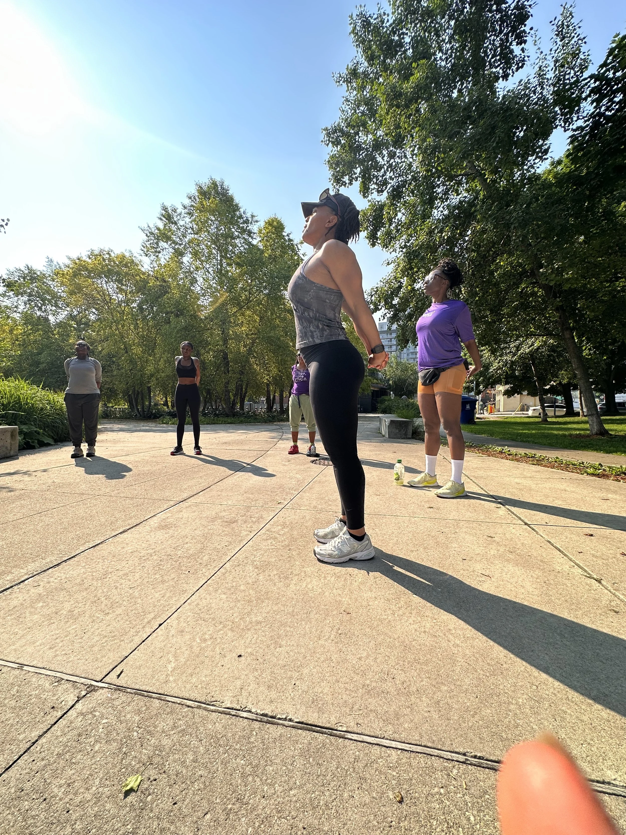 A group of five women exercising outdoors on a sunny day in a park, standing on a paved area surrounded by trees.