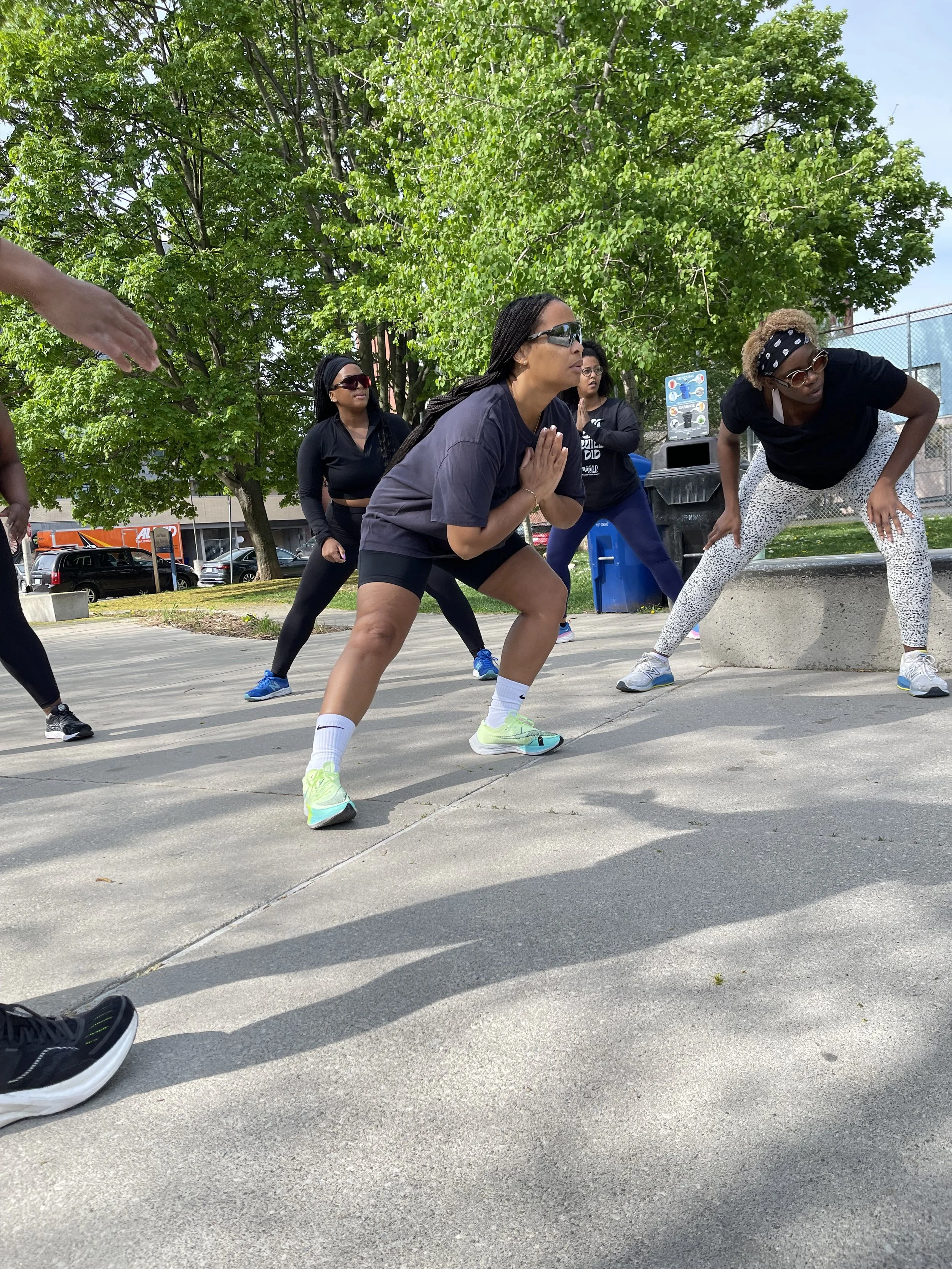 A group of women exercising outdoors in a park, performing different workout poses on a sunny day with green trees and blue sky in the background.