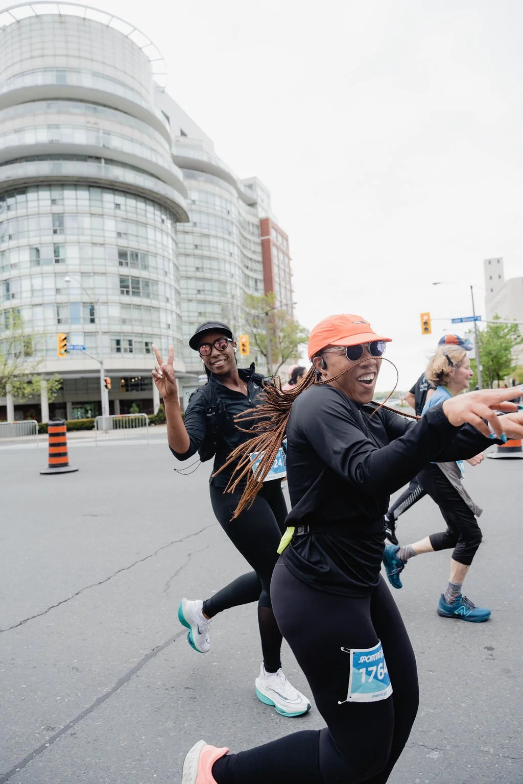 Two women running in a marathon, smiling and making peace signs, with a cityscape and traffic lights in the background.