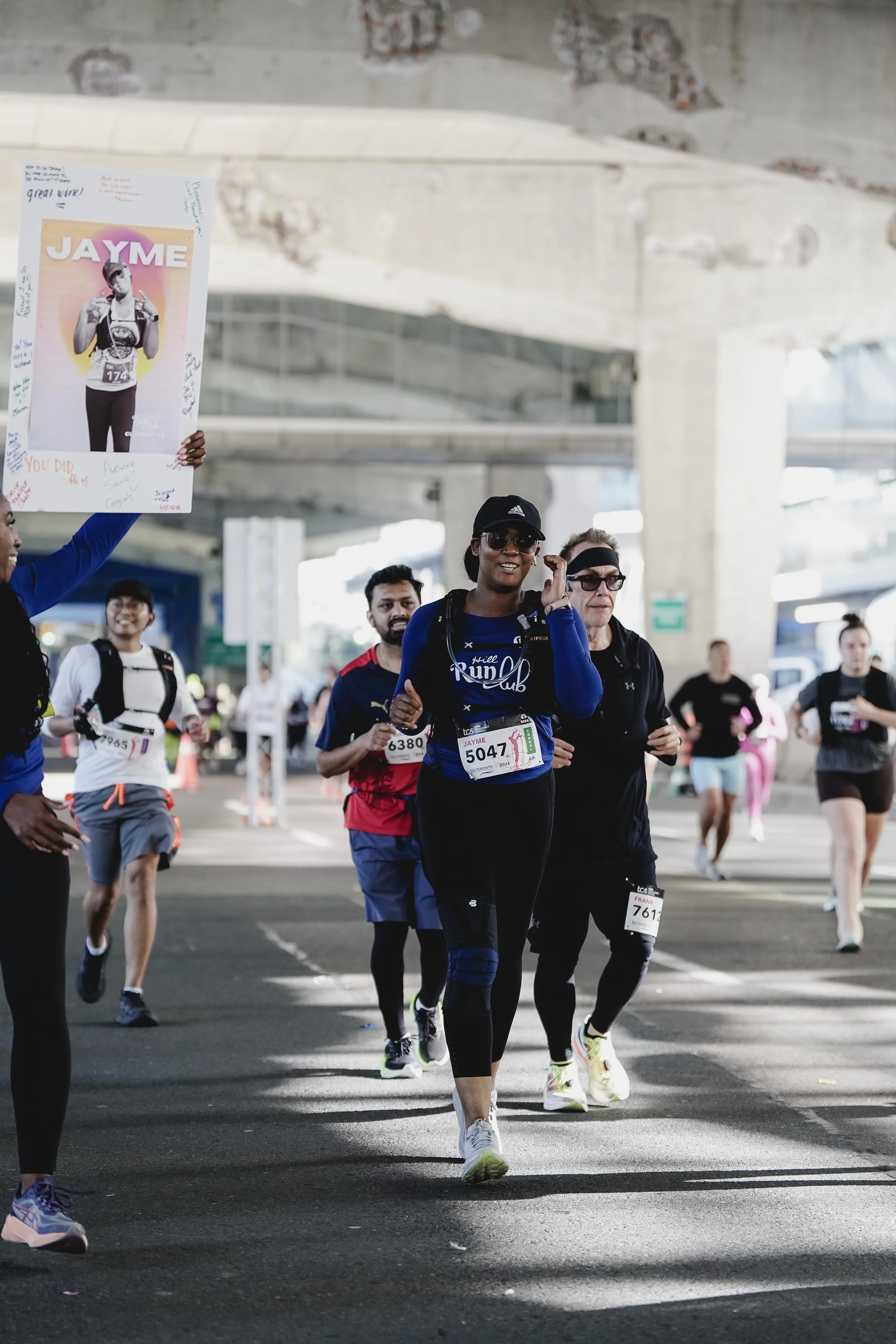 People participating in a marathon race under an overpass, with some holding signs supporting a runner named Jayme.