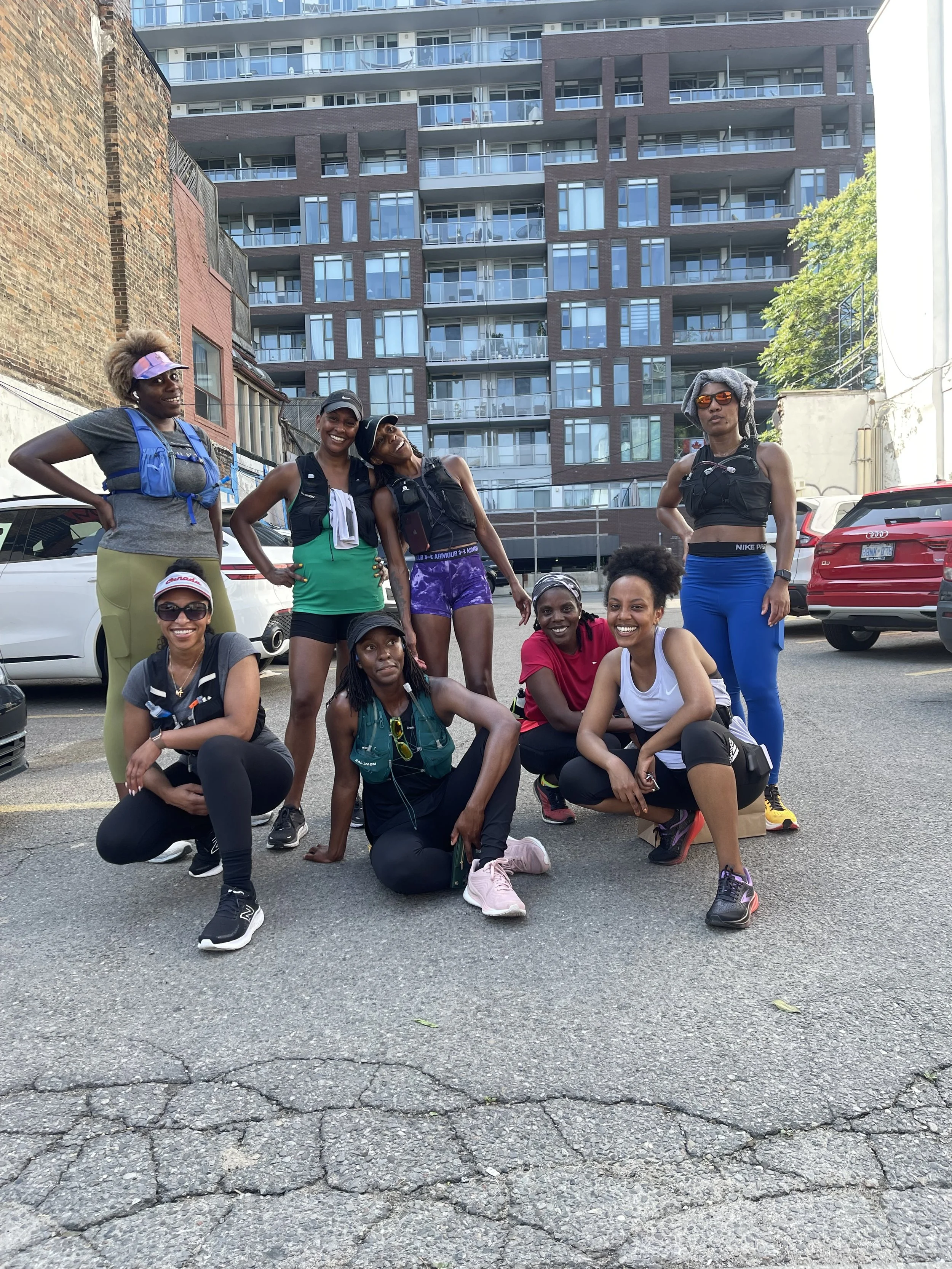 Group of nine women in athletic and casual clothing posing outdoors, with a parking lot and modern apartment buildings in the background.