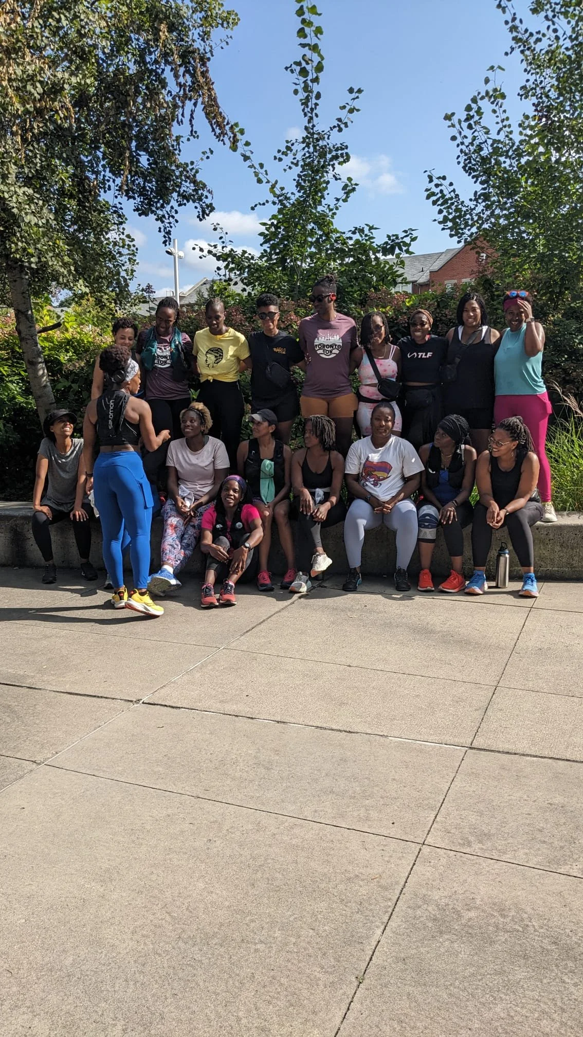 Group of women and girls outdoors on a sunny day, some sitting on a low wall and others standing behind them, surrounded by trees and bushes.