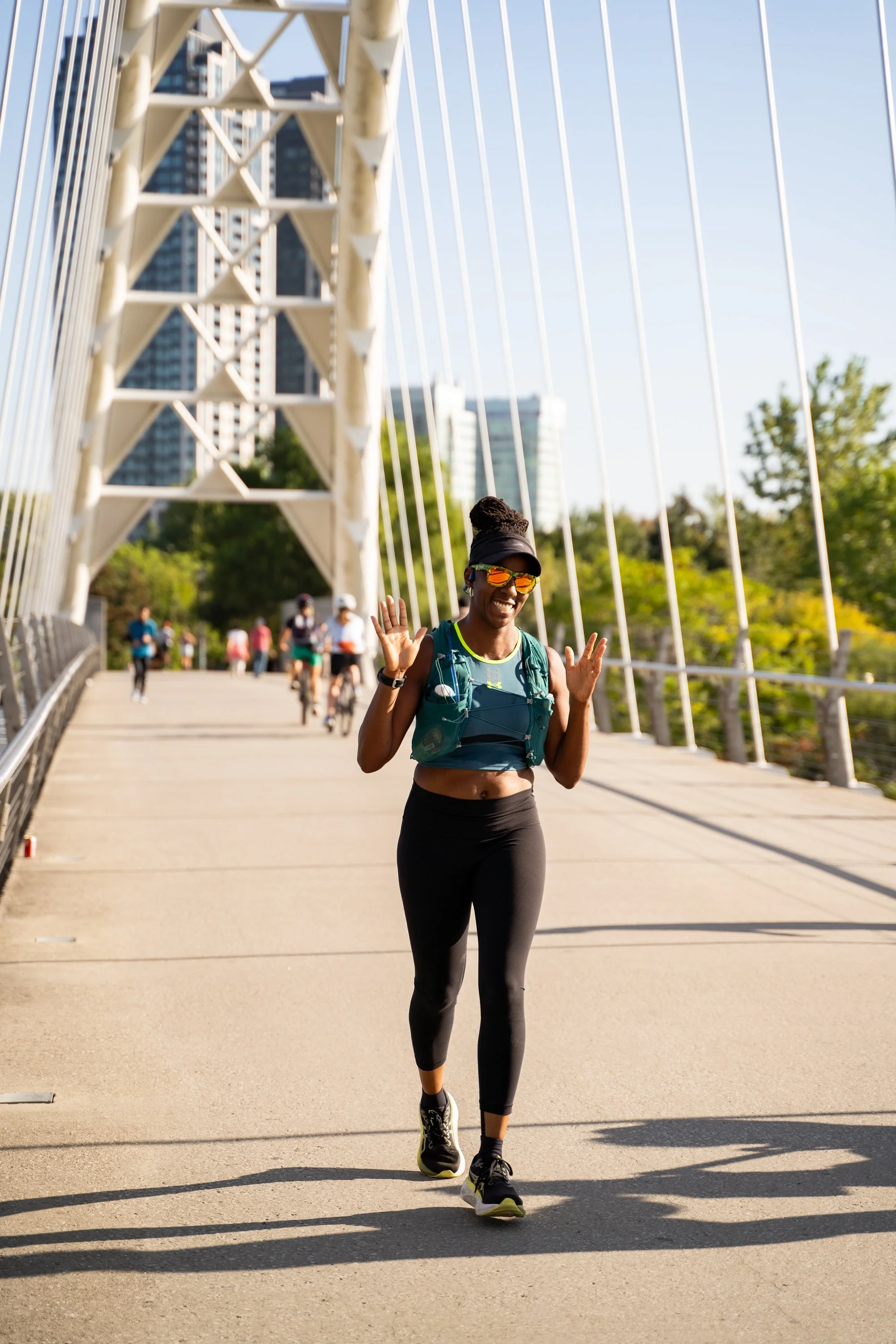 A woman running on a bridge, smiling and waving, with other runners in the background, in an outdoor urban park setting.