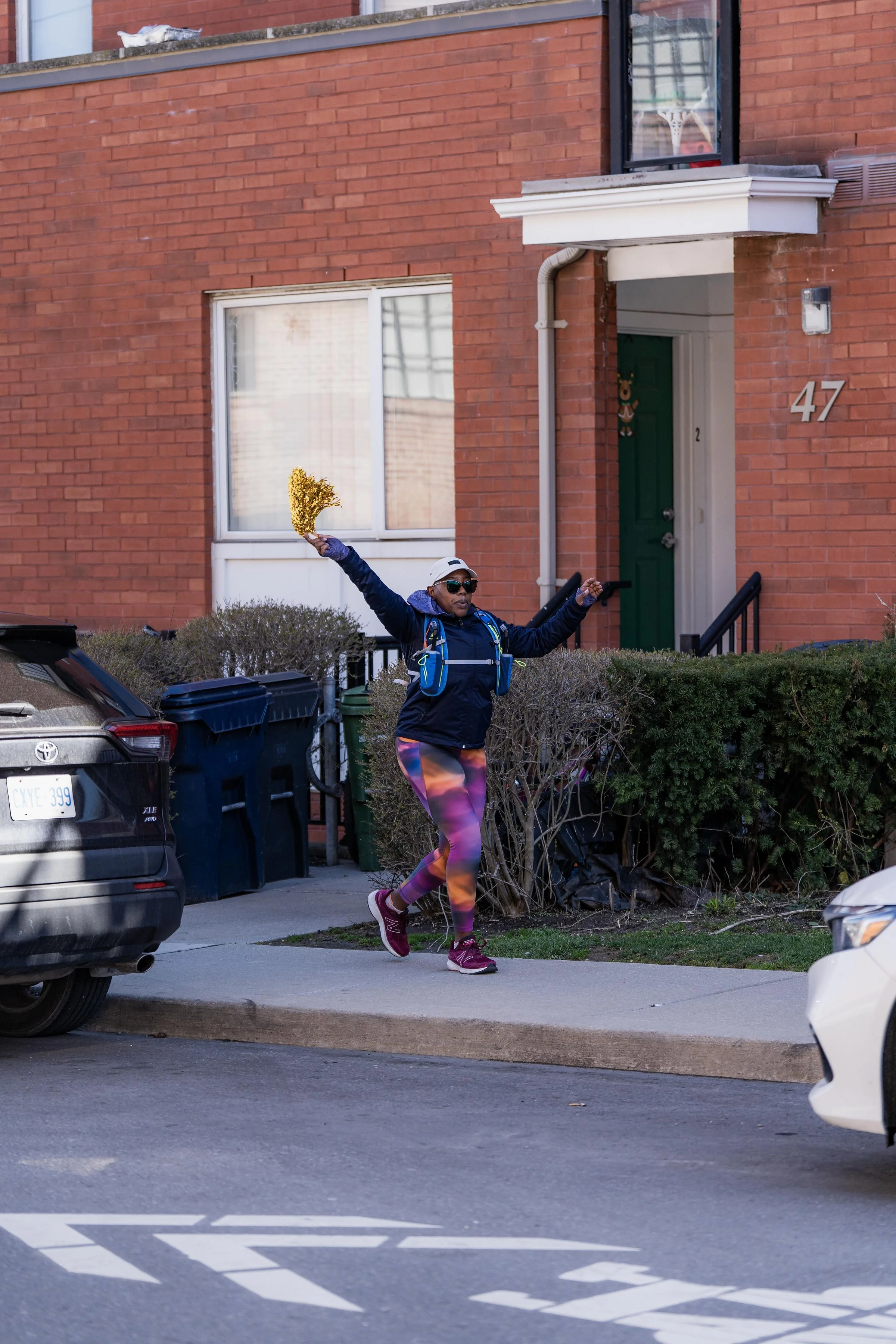 Person jogging on sidewalk near brick apartment building, holding a golden pom-pom, wearing colorful leggings, sunglasses, and a navy jacket, with a backpack and cap.