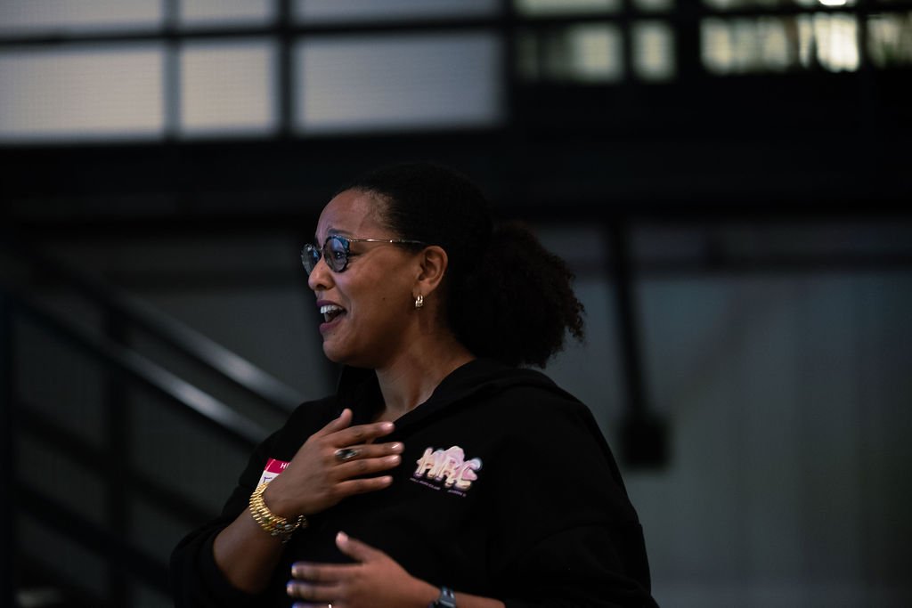 A woman wearing sunglasses and jewelry smiling and placing her right hand on her chest in an indoor setting.