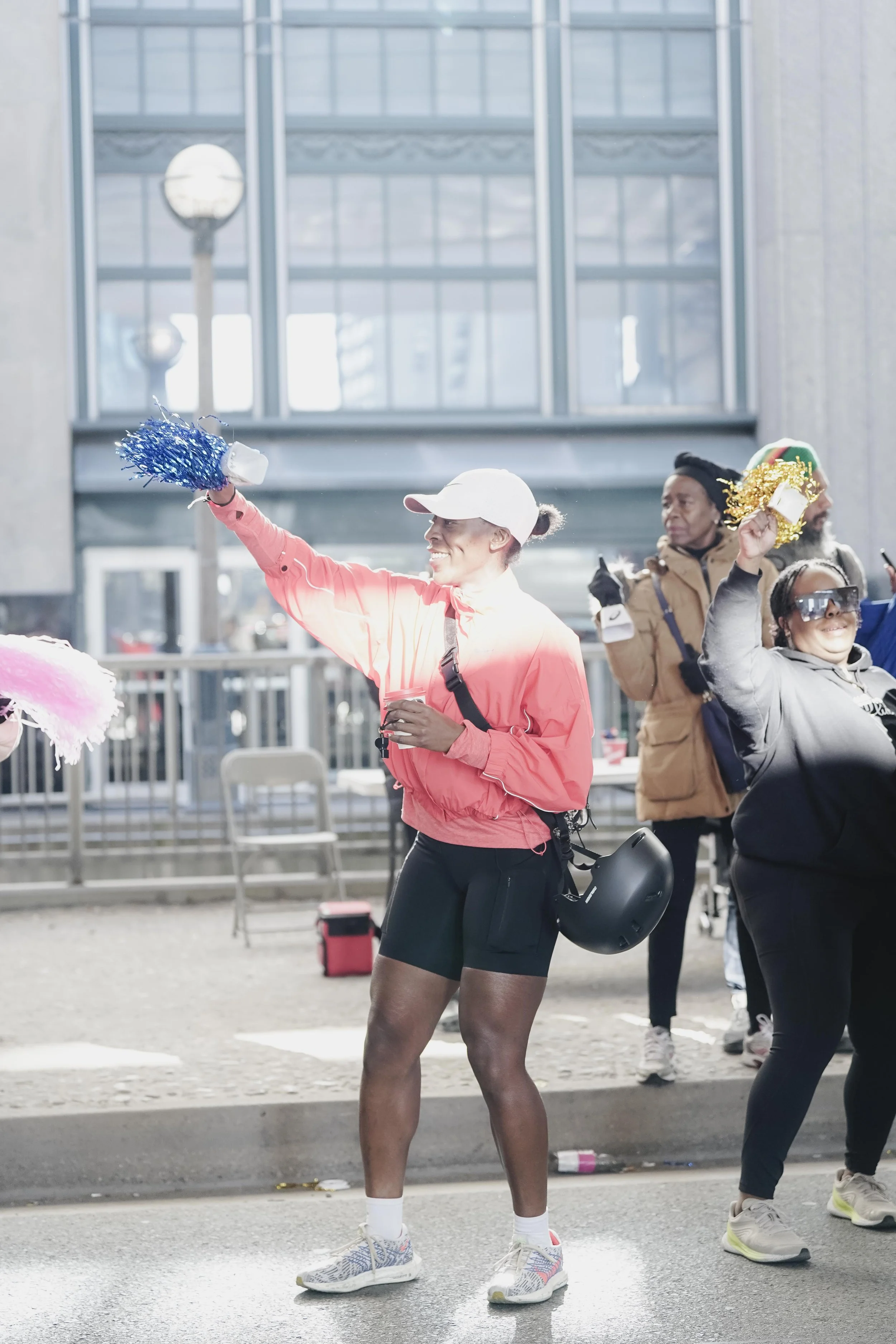 People celebrating at an outdoor event during daytime, some wearing athletic clothing and holding festive items, surrounded by urban buildings.