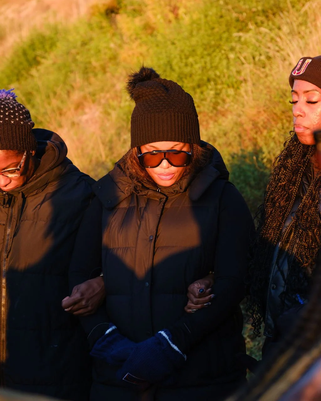 Three women standing outdoors wearing winter clothing and knit hats during sunset or sunrise.