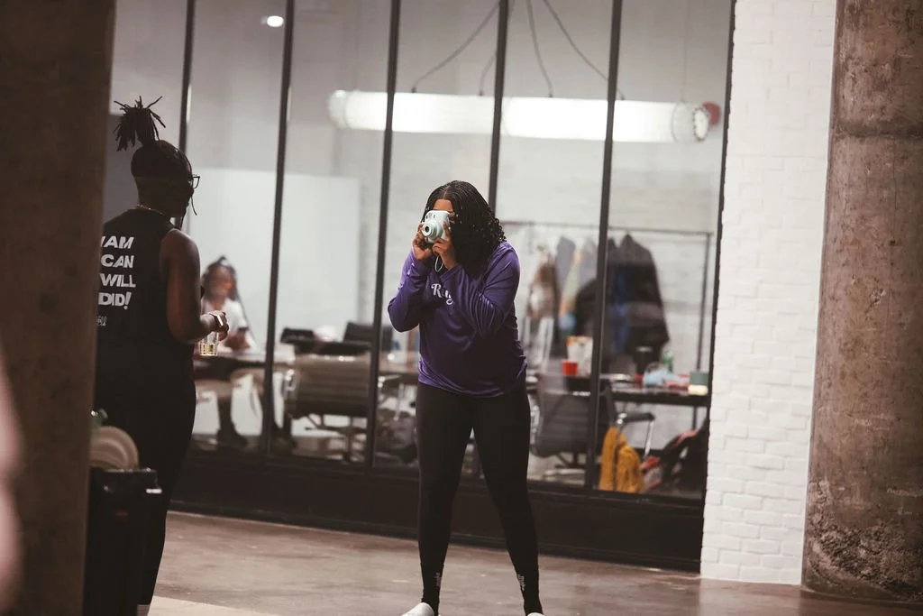 A woman taking a photo with a camera in an industrial-style office or studio space reflected in a large glass wall. Other people are working or socializing in the background.