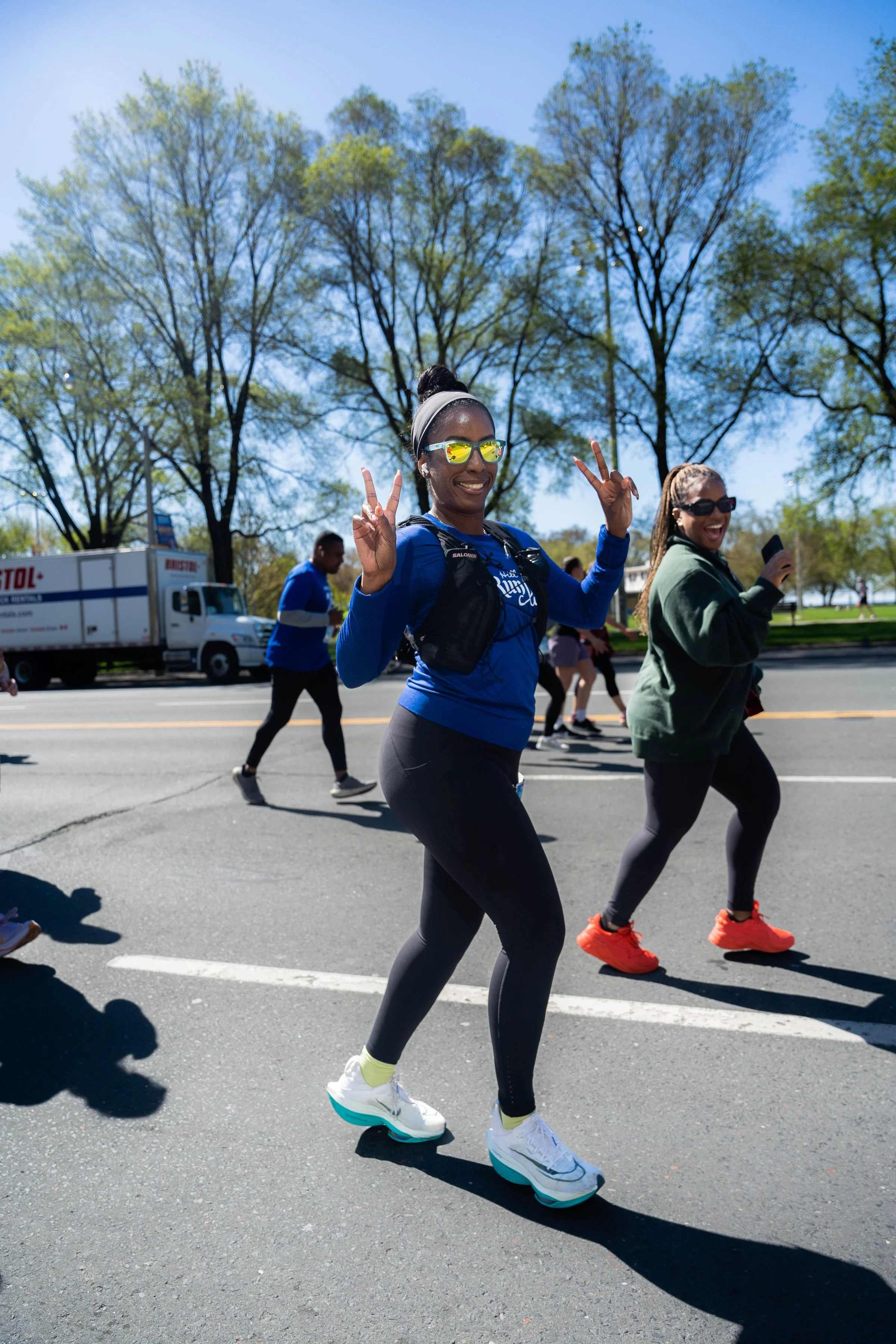 Women running in a race, one making peace signs and smiling, on a sunny day with trees in the background.
