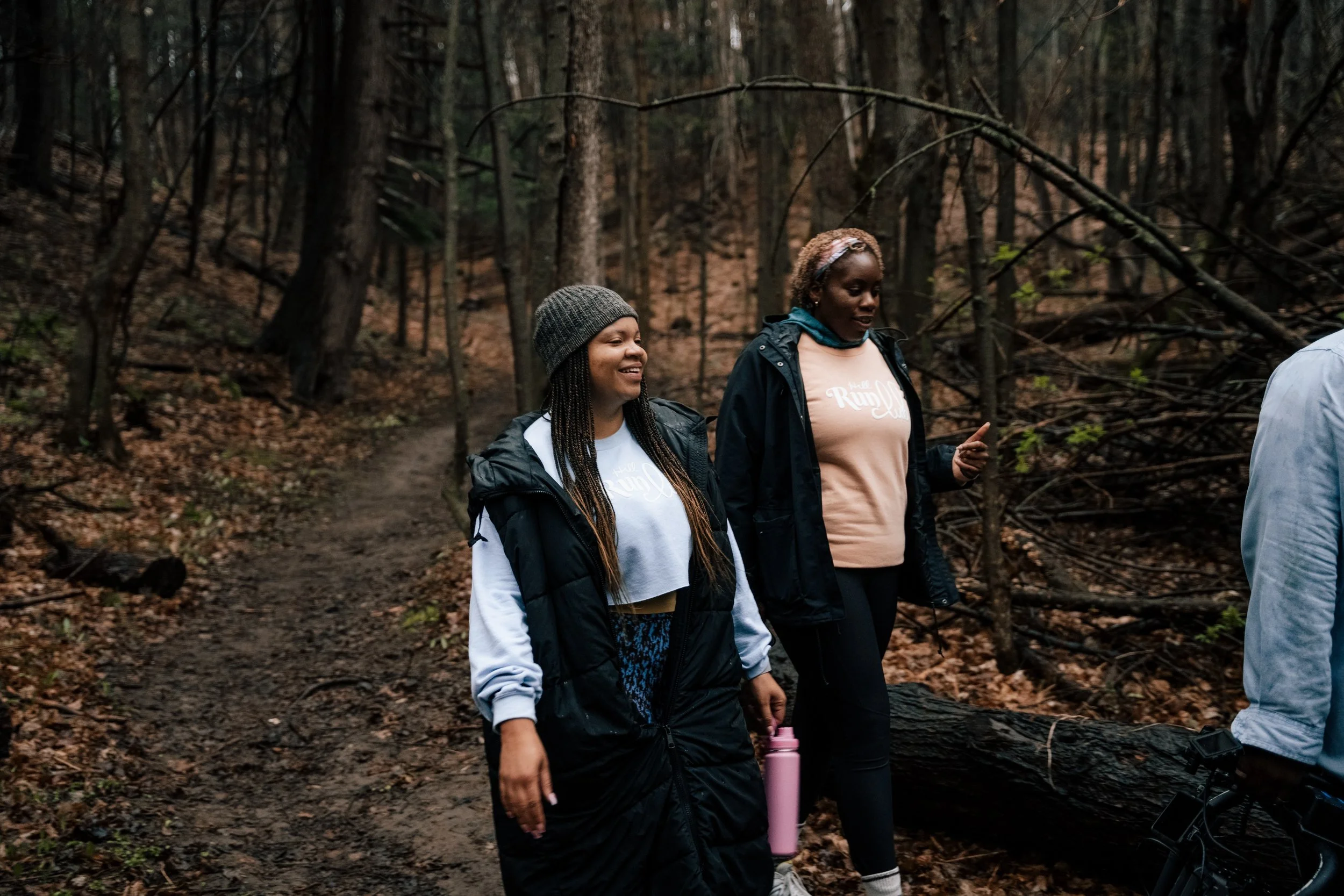 Two women walking along a forest trail surrounded by trees and fallen leaves on an overcast day.
