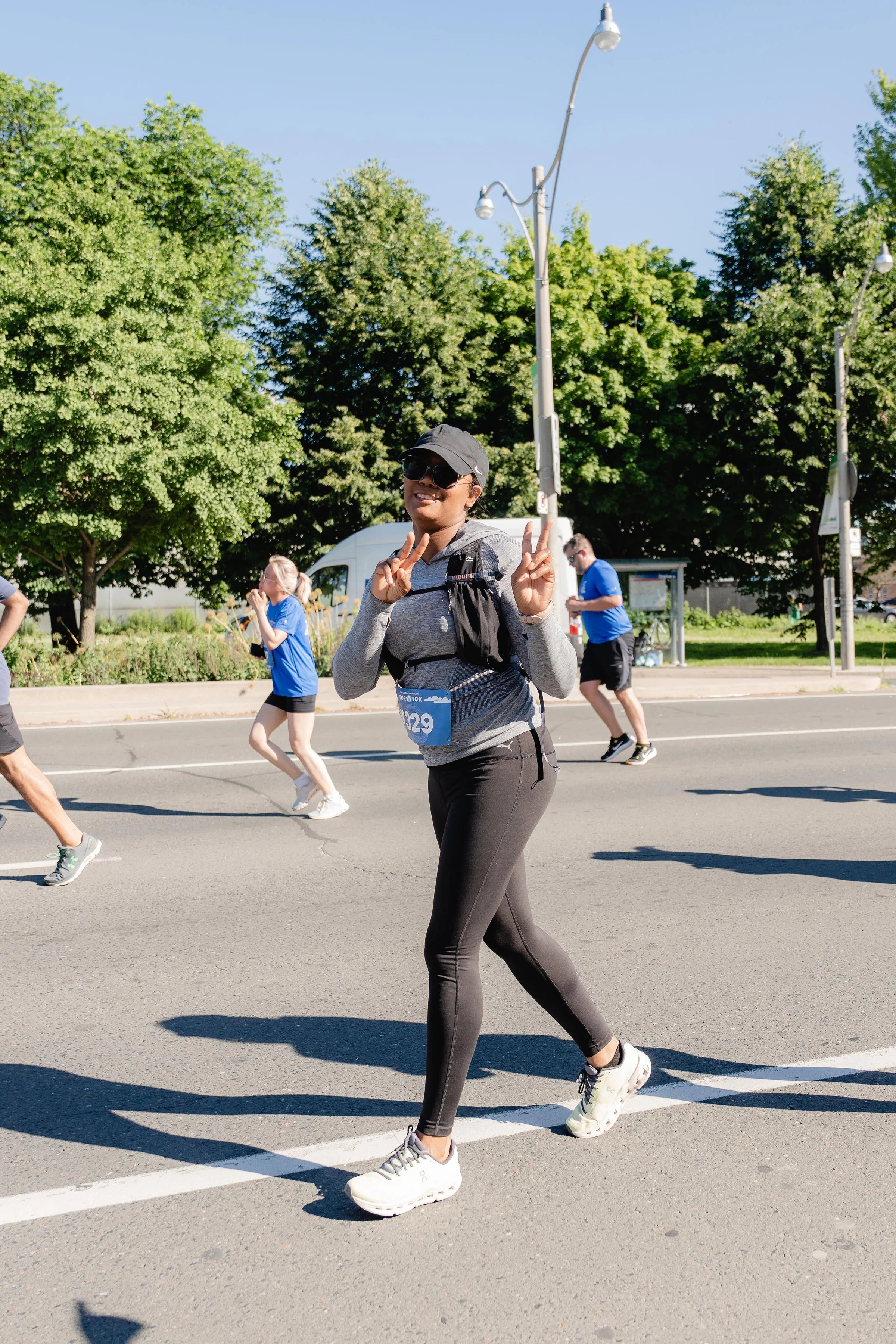 A woman smiling and making peace signs while running in a marathon on a sunny day, with other runners and green trees in the background.
