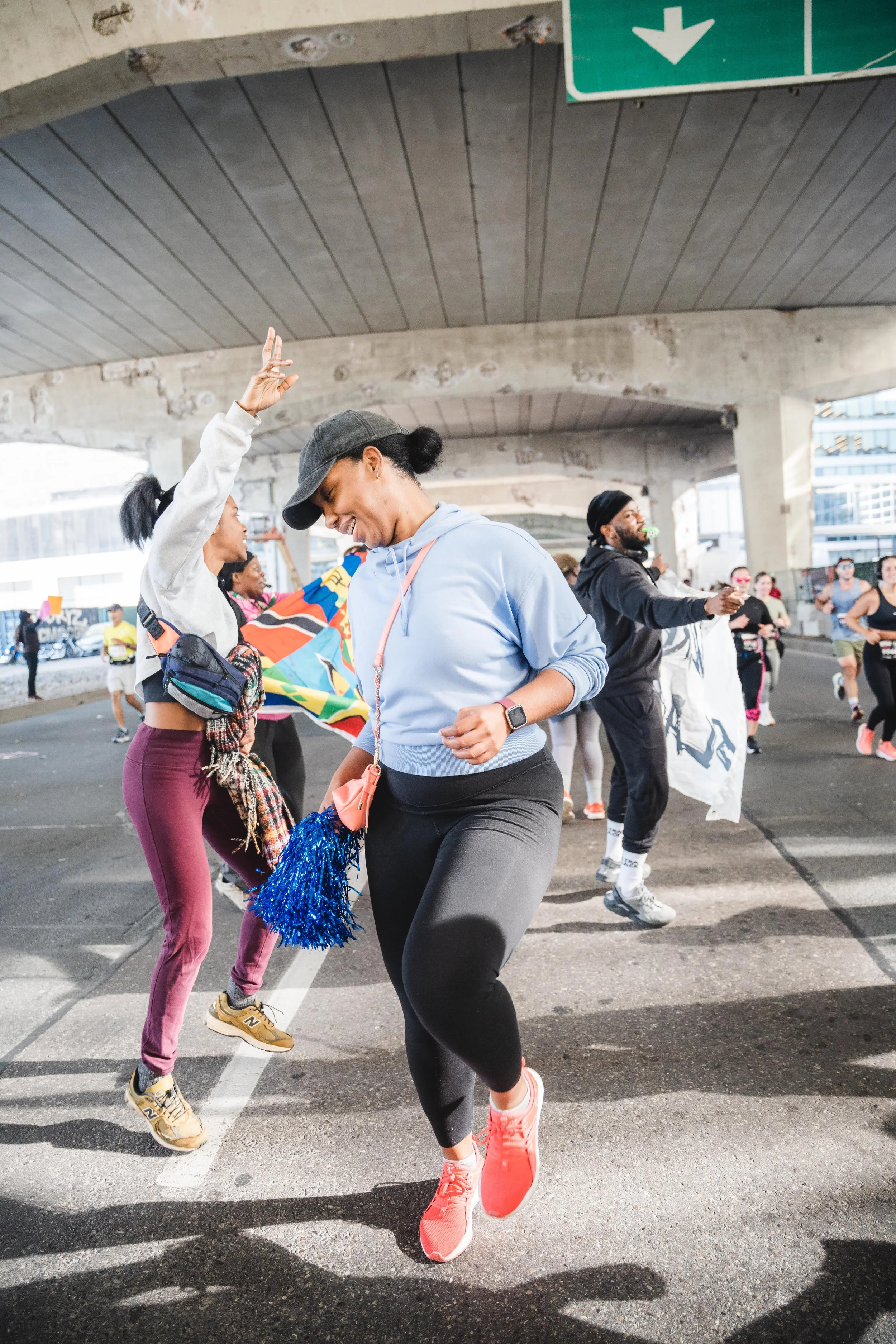 People dancing and celebrating under a bridge during an outdoor event or race, with some holding flags and bags.