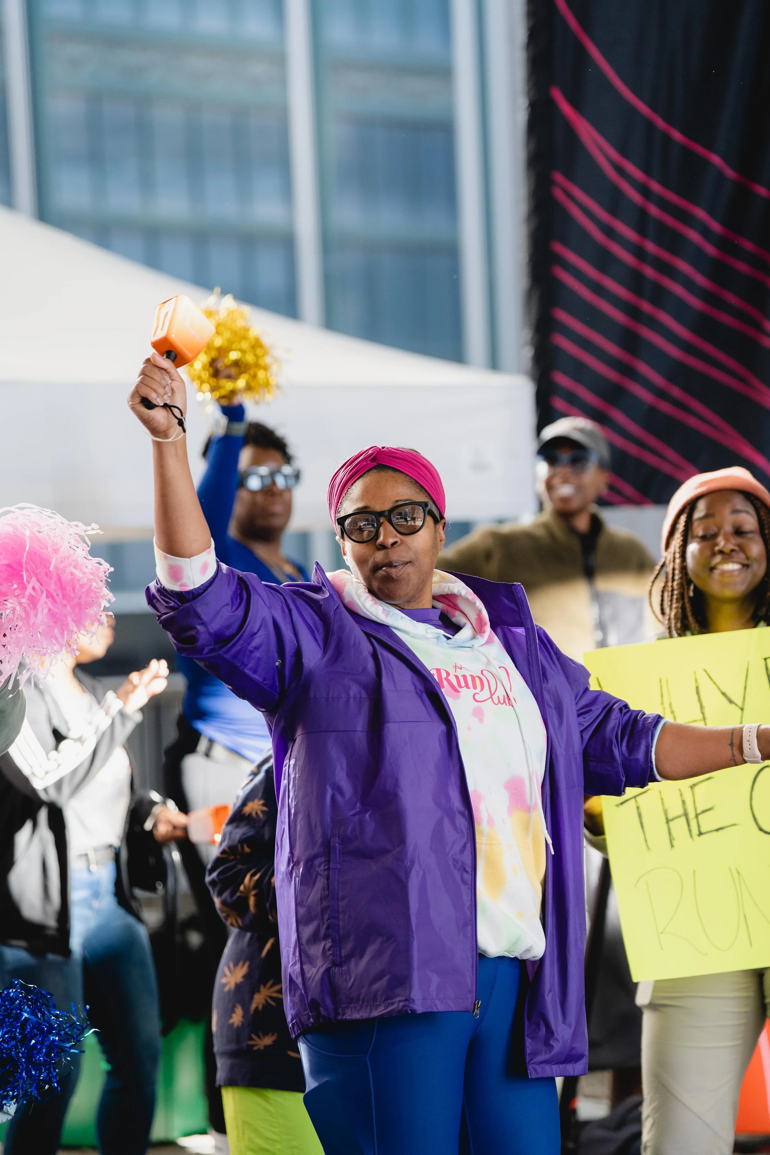 A woman with glasses and a pink headband holding a plastic hammer and a yellow sign at a street protest or rally.