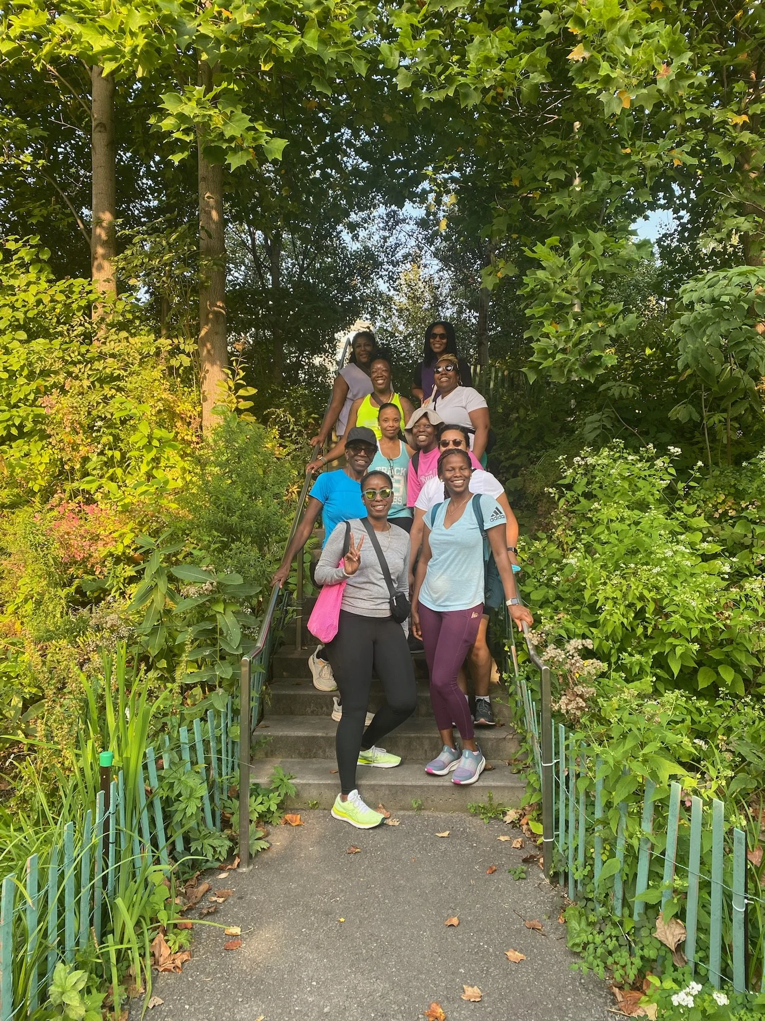 Group of smiling women and girls standing on steps surrounded by lush green trees and bushes during daytime, enjoying a walk outdoors.