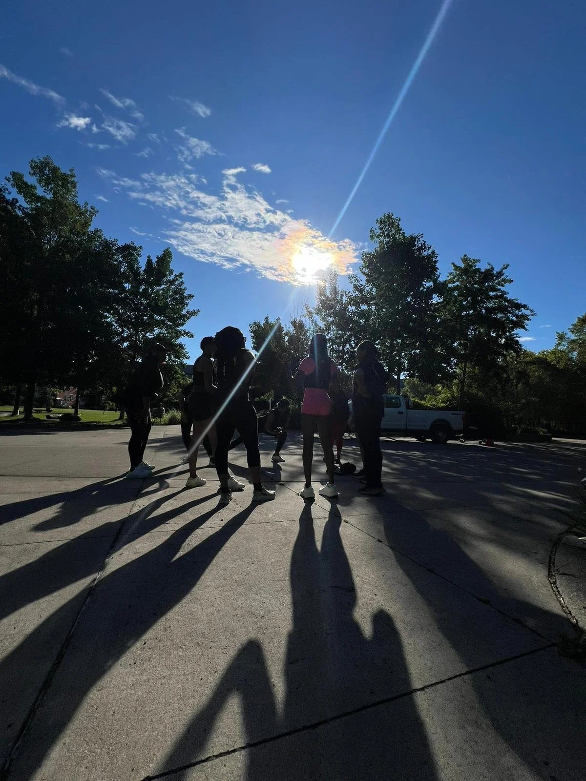 Group of women gather outdoors on a sunny day, shadows cast on pavement, trees and a truck visible in background.