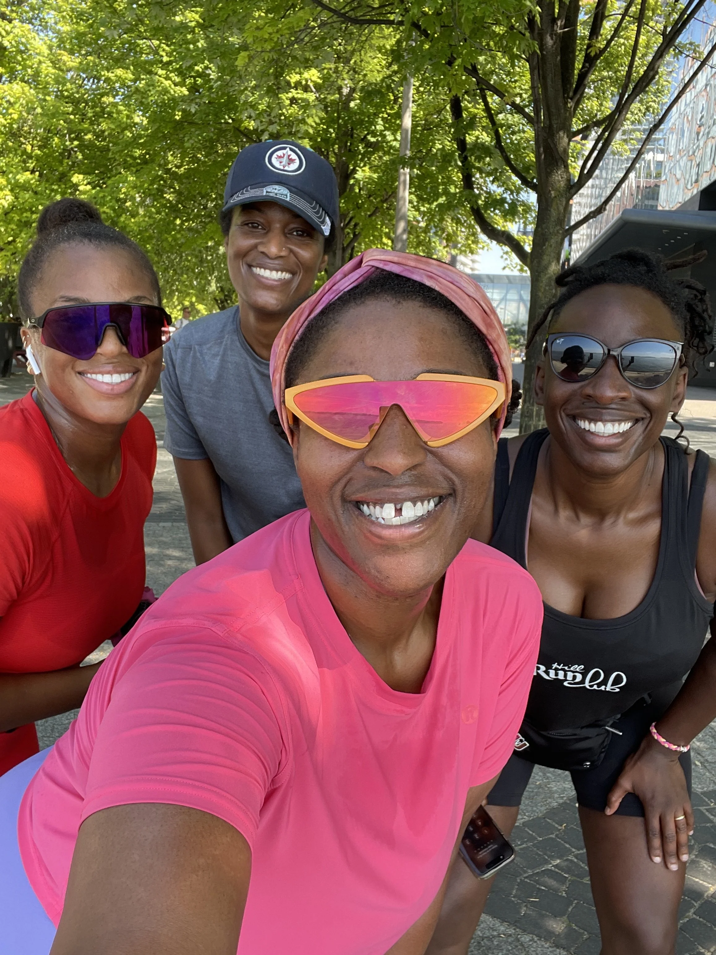 A group of four women smiling for a selfie outdoors on a sunny day, with green trees and a modern building in the background. They are wearing athletic clothing and sunglasses.