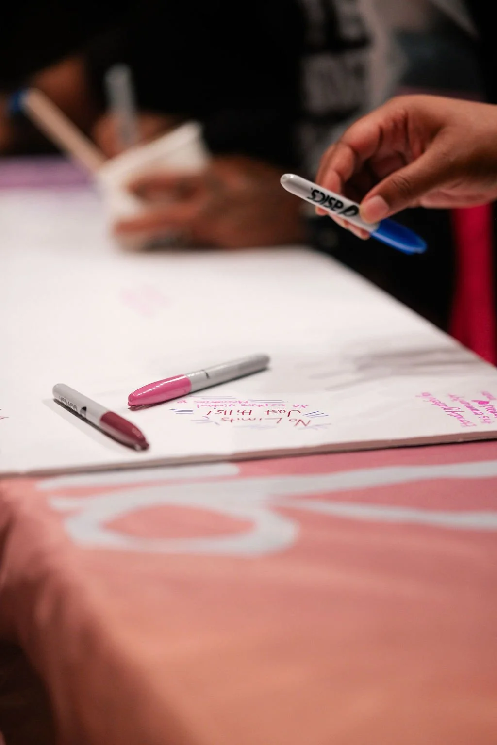 A person is writing on a piece of paper with pink and blue markers, and there are additional markers on the table.