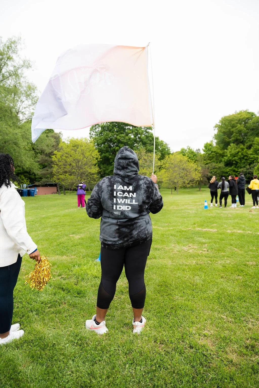 Person in black hoodie with motivational words standing on grass field holding a white flag during outdoor event with other people in the background.