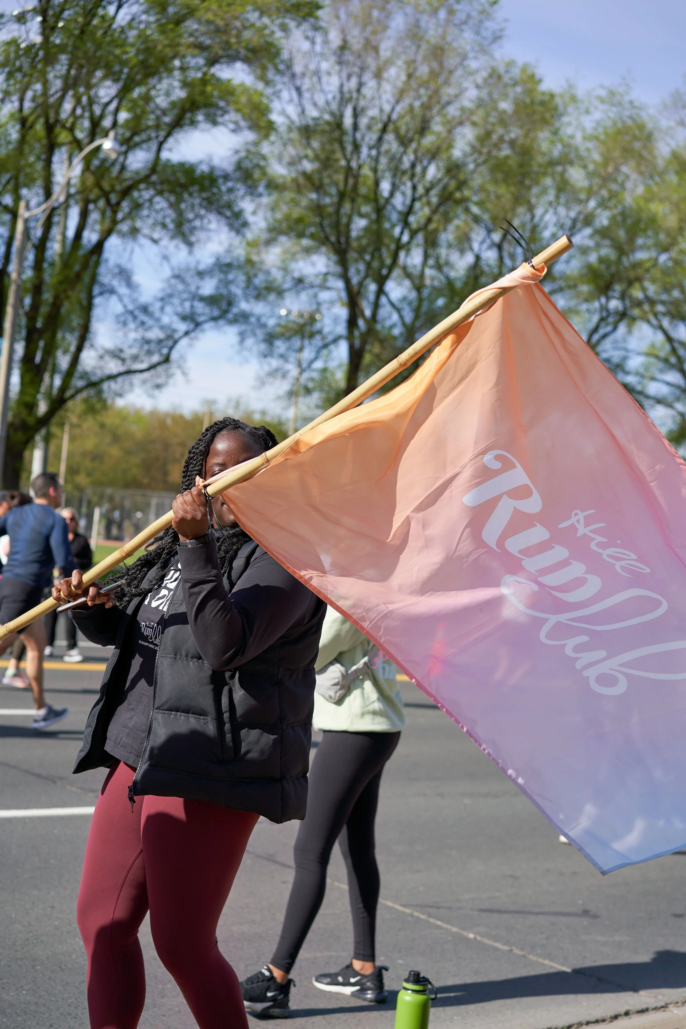 A woman holding a peach-colored flag with white cursive that says 'Kille Tranquil' during an outdoor event with trees and multiple people in the background.