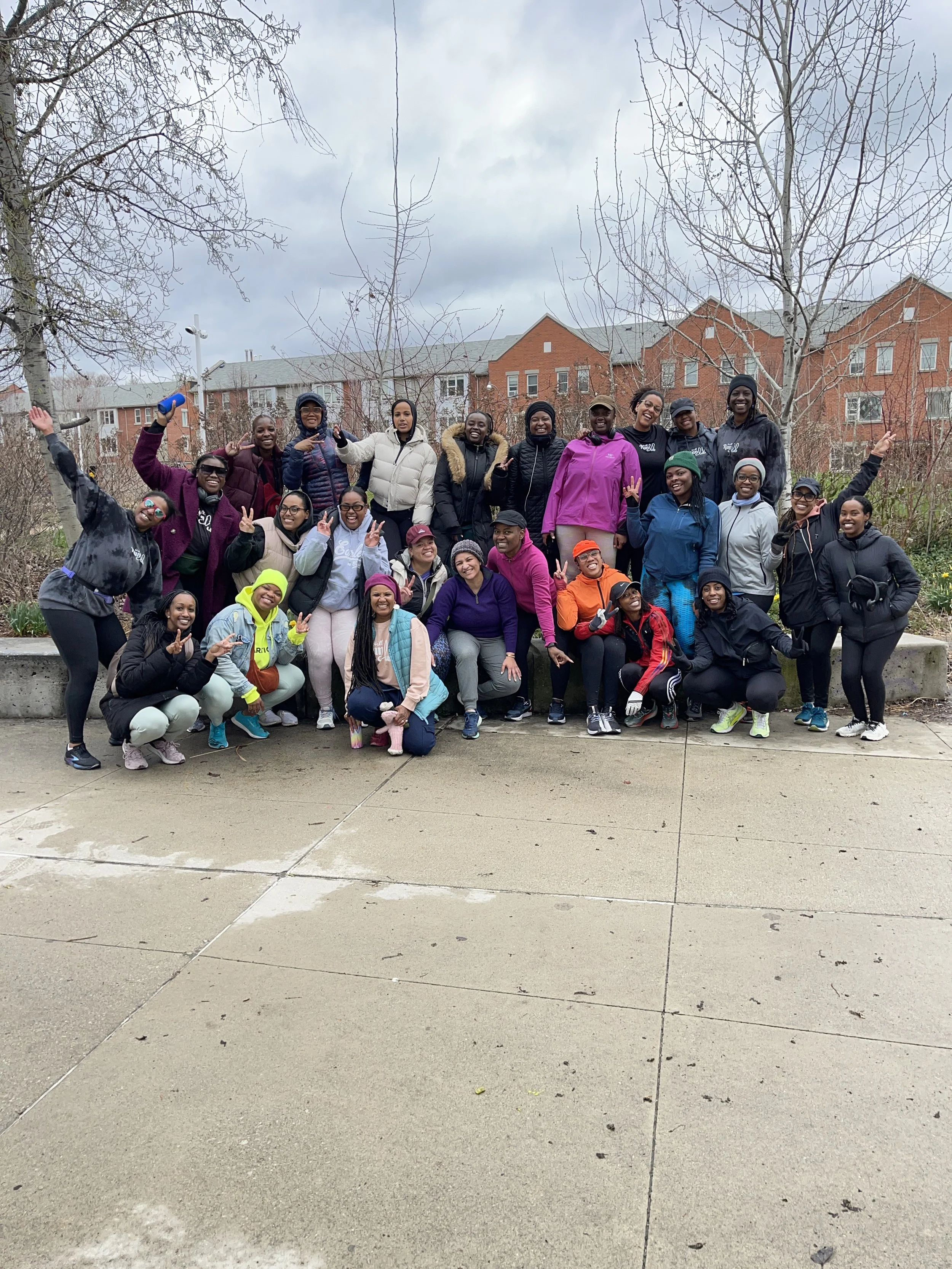 A group of diverse women and girls in athletic clothing and jackets poses outdoors on a sidewalk in front of trees and residential buildings. They are smiling, making peace signs, and raising their arms.