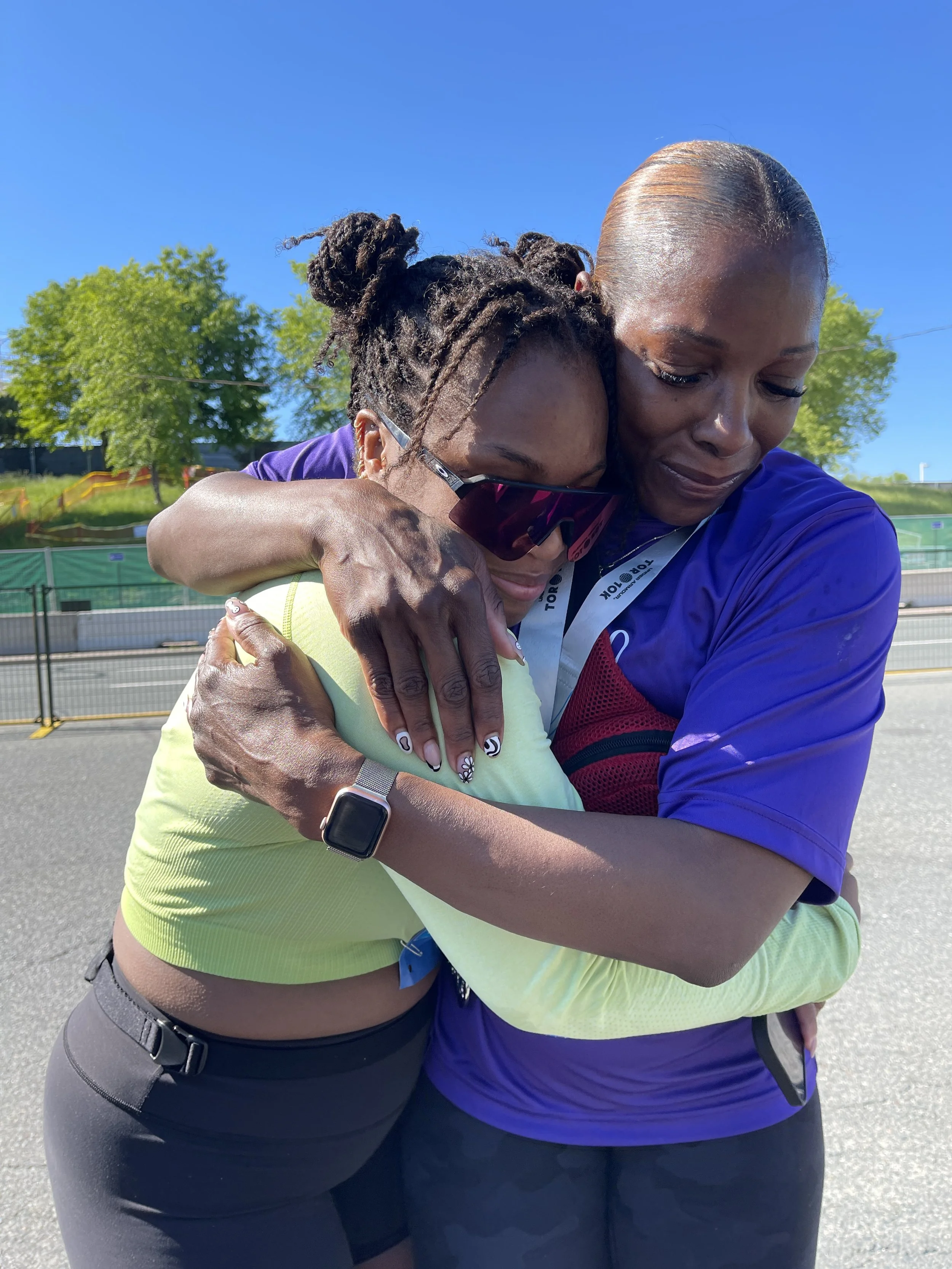 Two women hugging outdoors on a sunny day, one wearing sunglasses and the other wearing a blue athletic shirt.