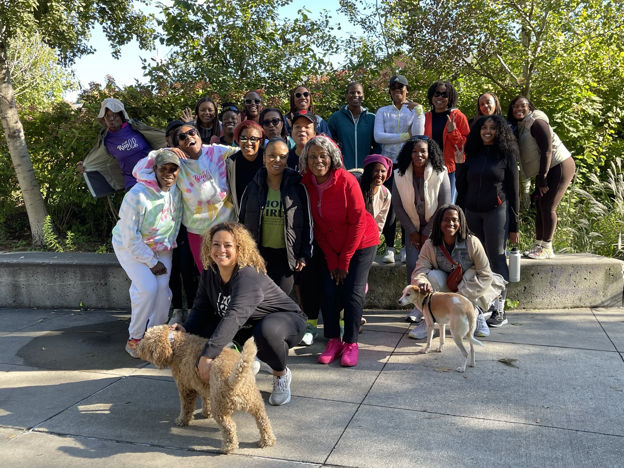 Group of women outdoors on a sunny day, posing for a photo with two dogs, in front of green trees and bushes.