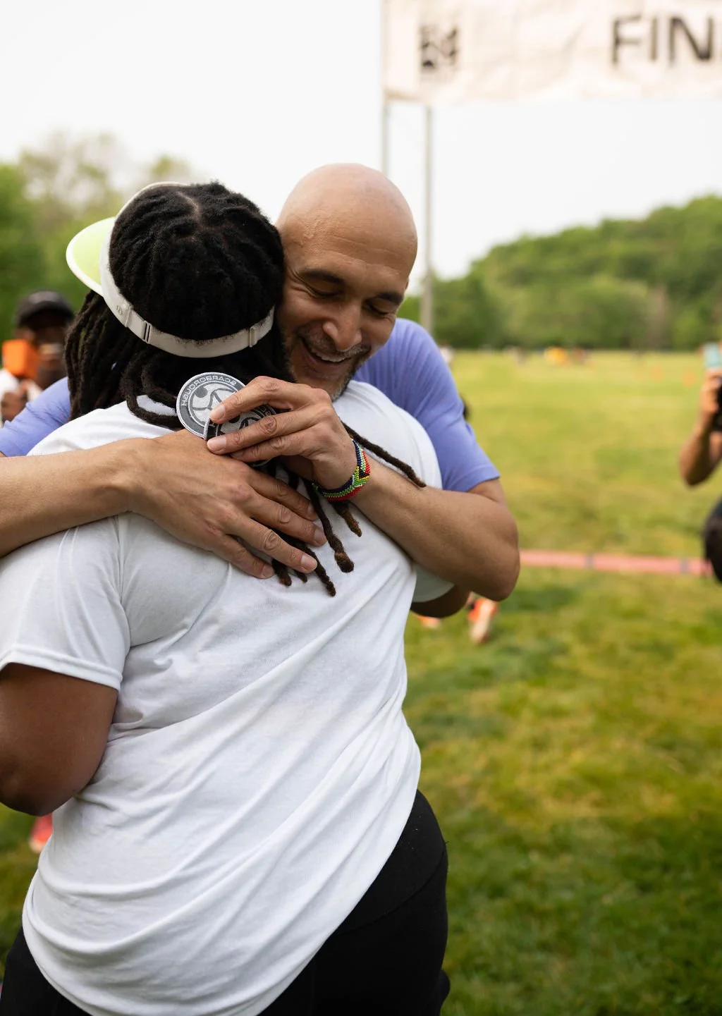 Two men hugging after a race finish, one with dreadlocks and wearing a white shirt, the other with a shaved head and wearing a blue shirt.