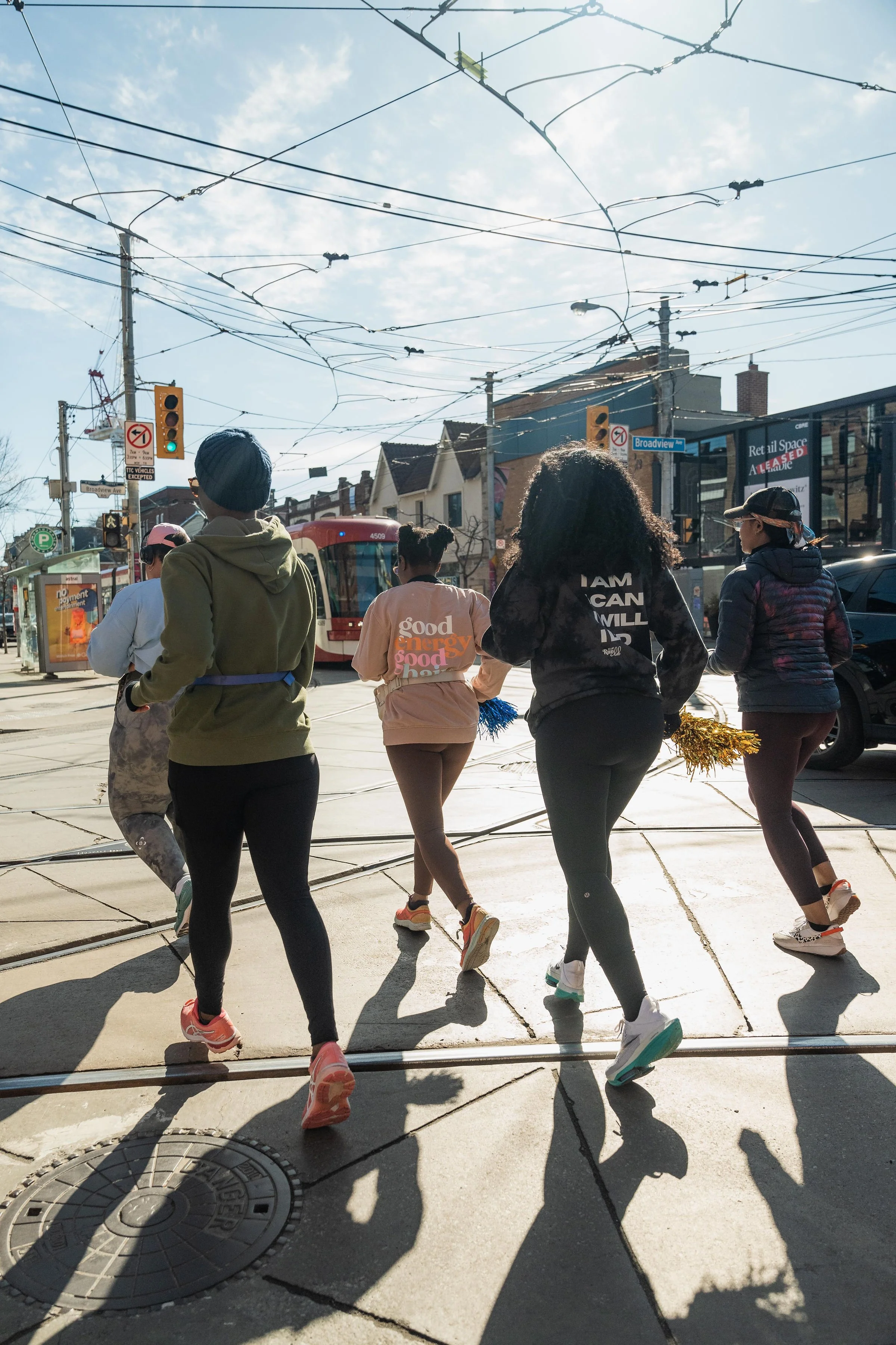 Group of women jogging through an urban intersection with tram tracks, overhead wires, traffic lights, and buildings in the background on a sunny day.
