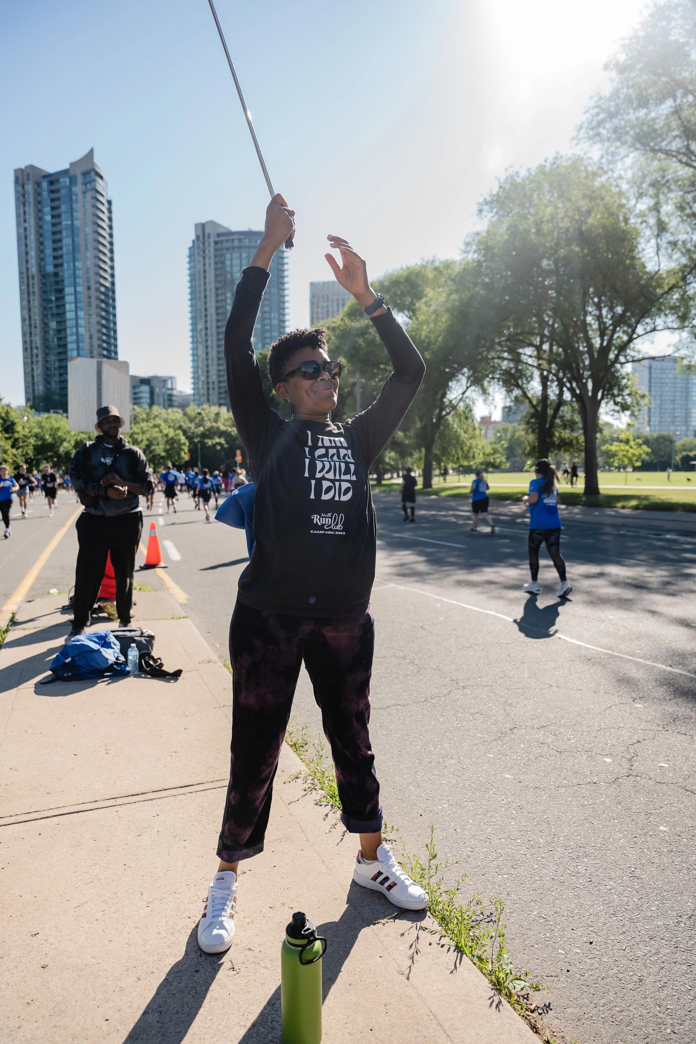 A woman standing on a sidewalk, lifting a jump rope above her head during a fitness event in an outdoor park with tall buildings in the background.