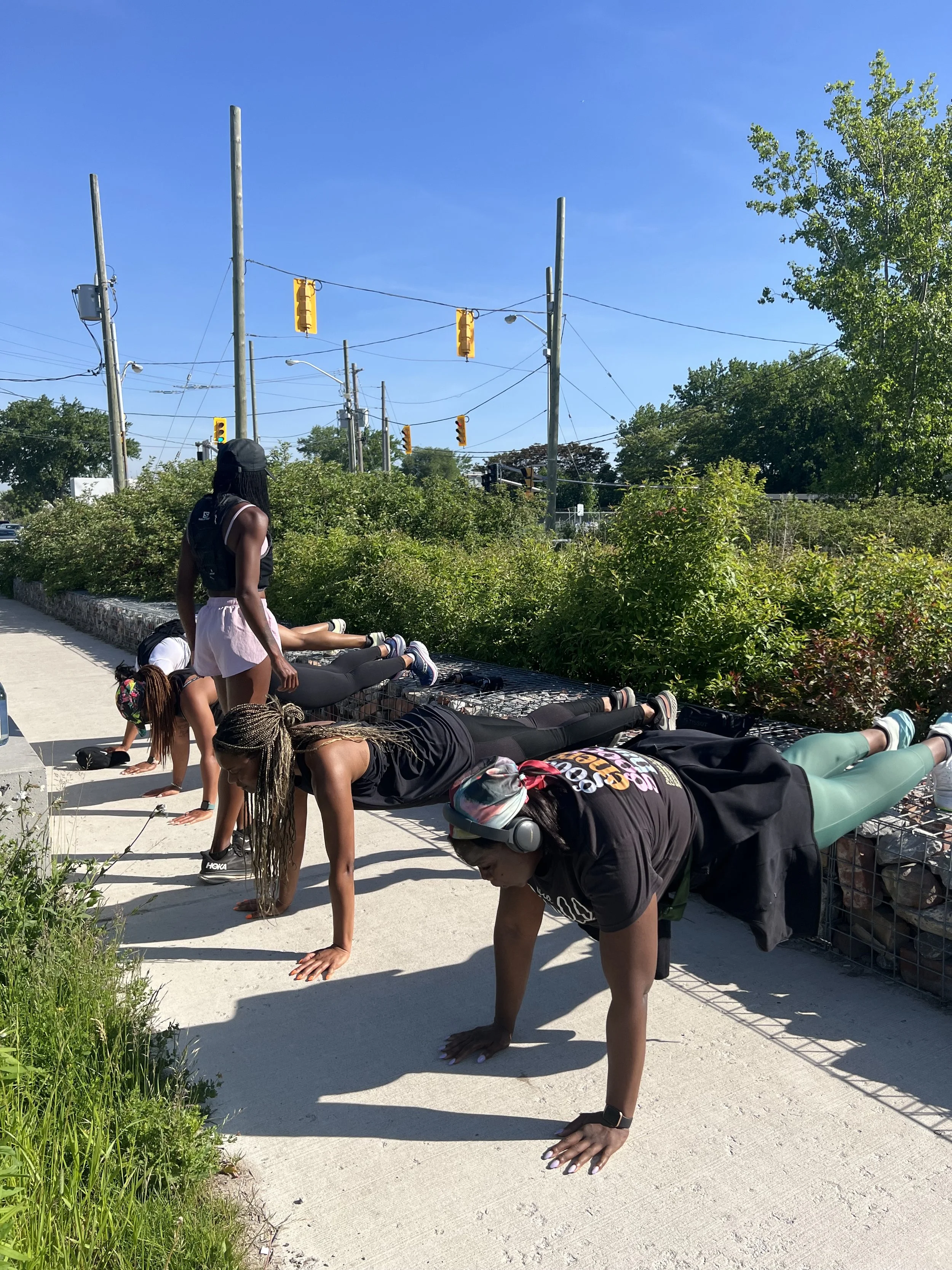Women doing push-ups outdoors on a sidewalk with green bushes and utility poles in the background.