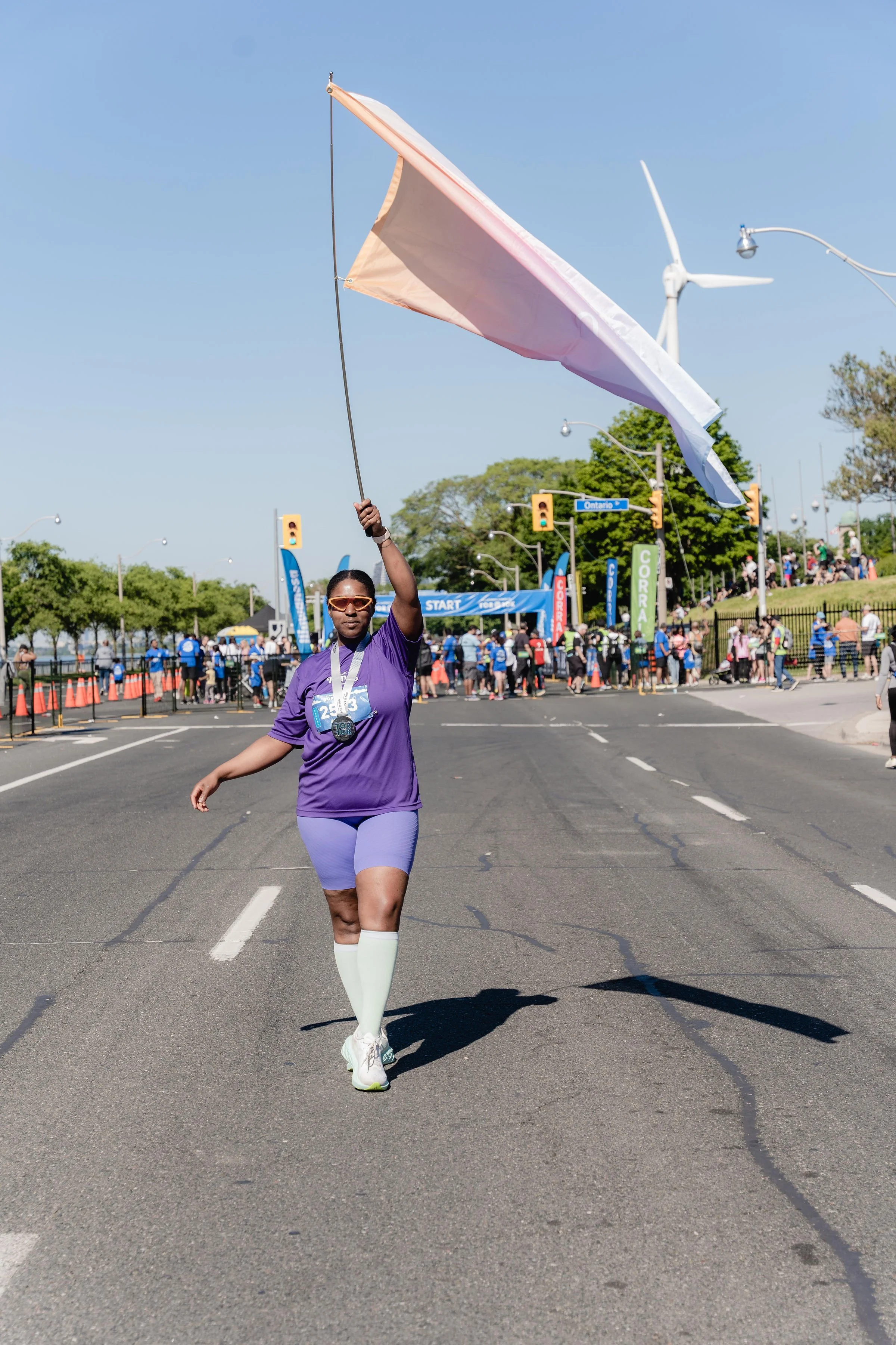 A woman in a purple athletic outfit is running and waving a white flag at a race event. She is wearing sunglasses, a medal, and has a race bib on her chest. There are spectators and race officials in the background, with banners and an inflatable sta