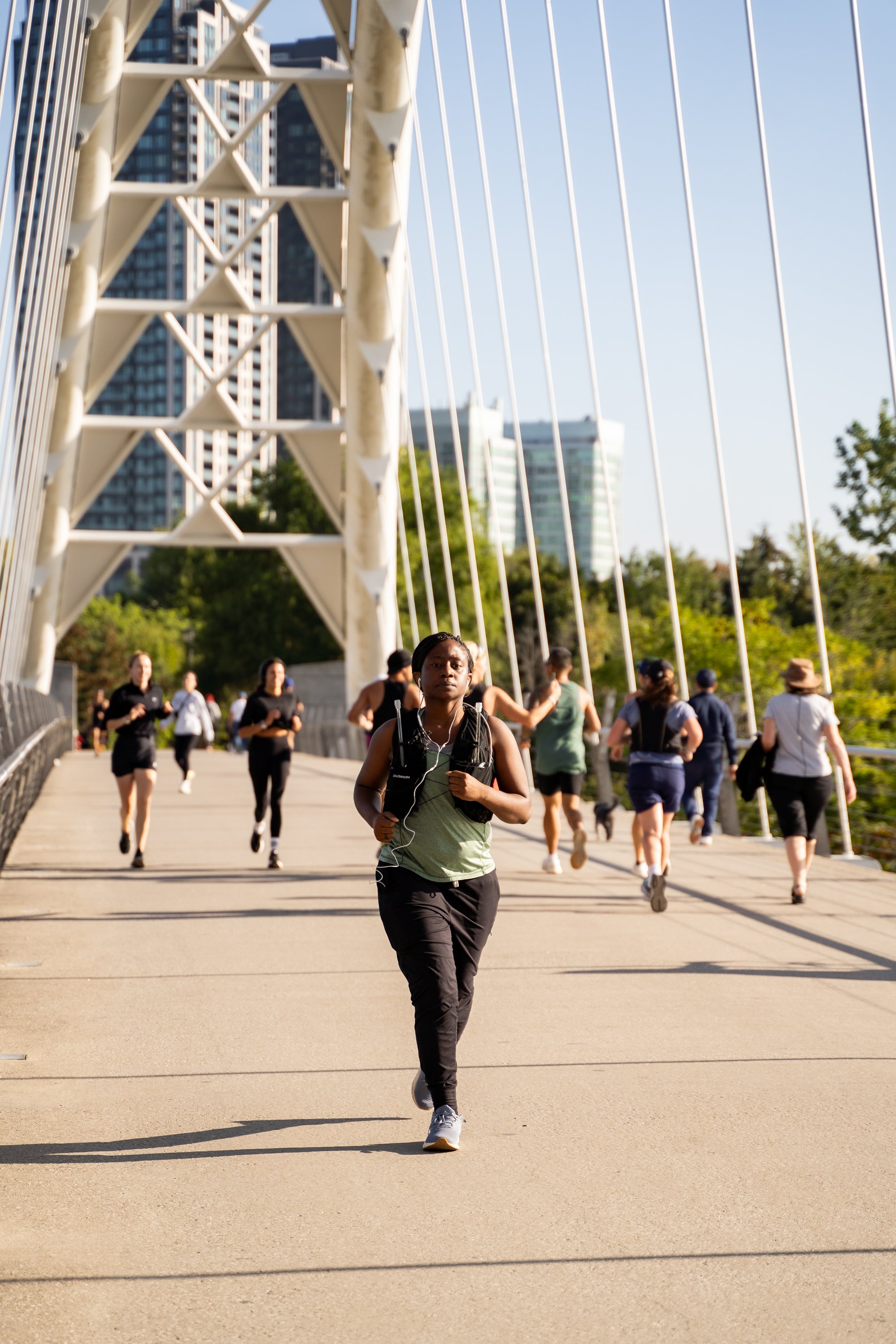 People jogging on a bridge in an urban park, with skyscrapers and trees in the background.