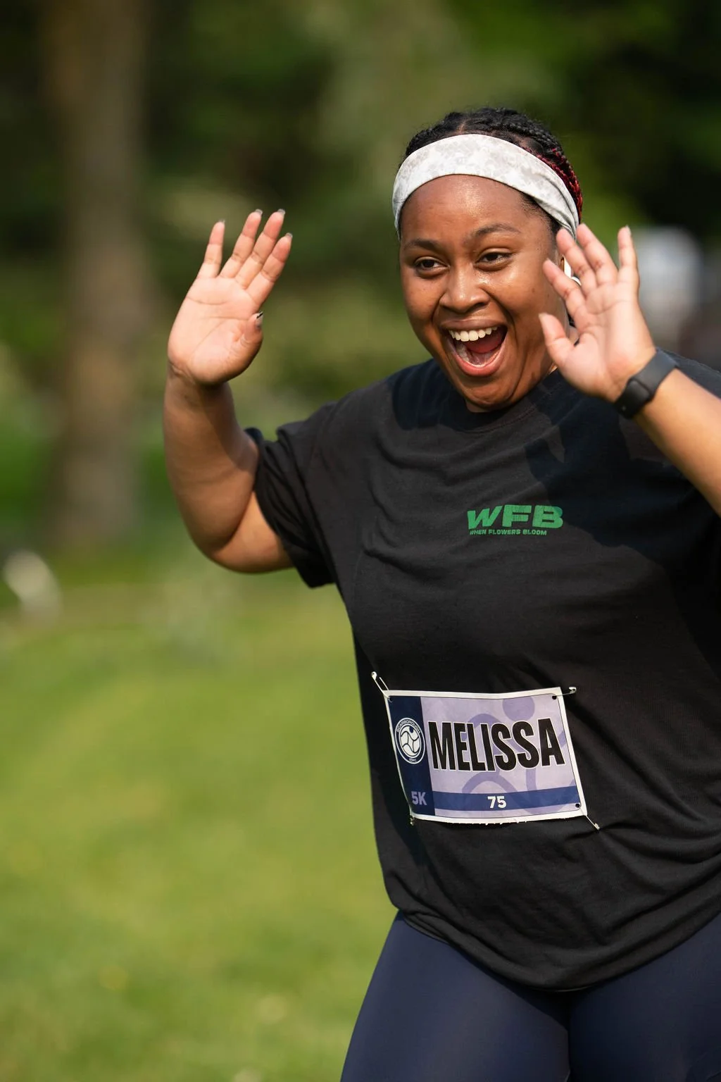 A woman with a white headband smiling and raising her hands in a celebratory gesture during a race, wearing a black shirt with a name tag that reads 'Melissa' and a green logo.