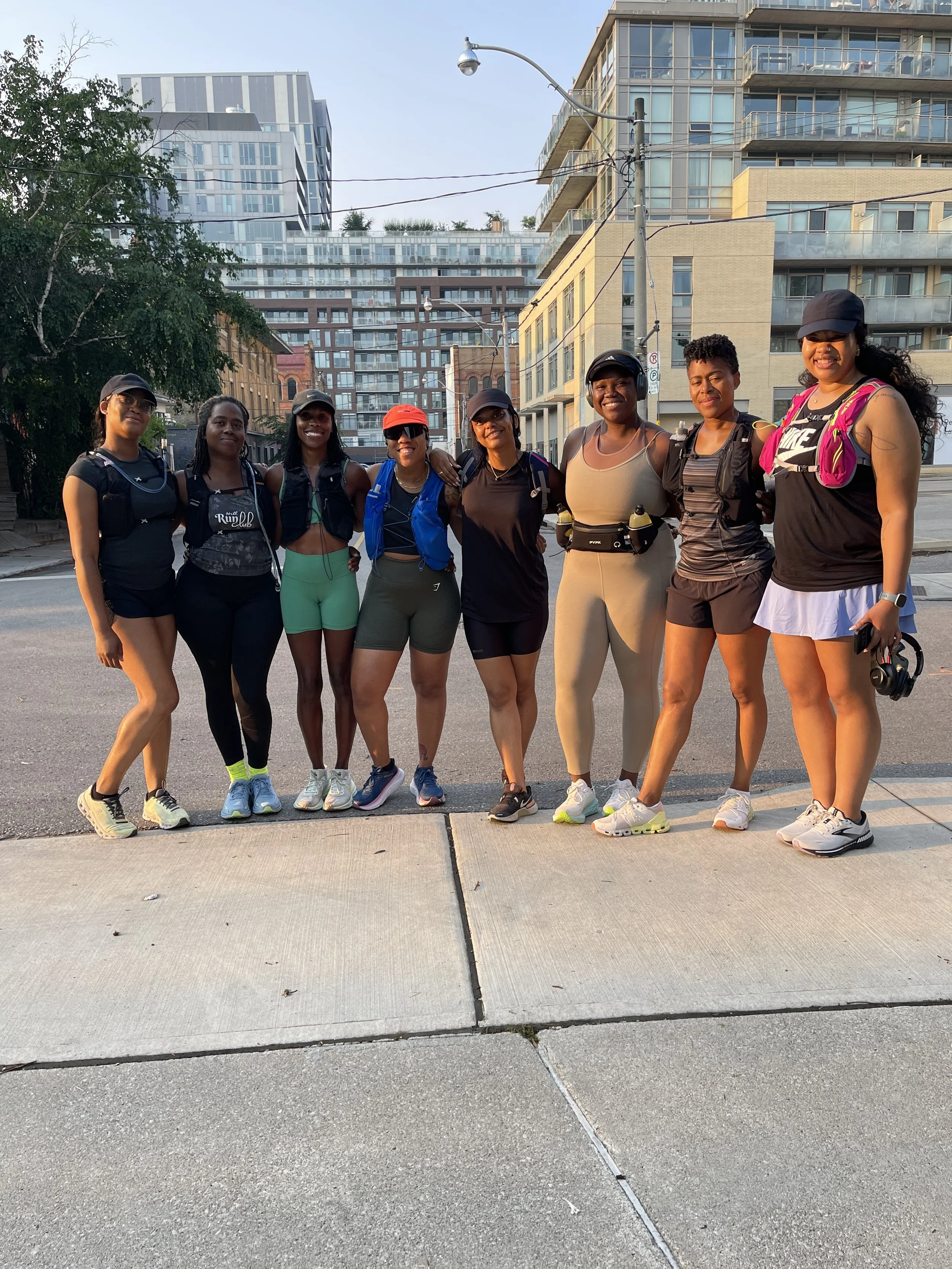 Group of nine women dressed in athletic gear, standing close together on a city sidewalk, smiling at the camera with urban buildings in the background.