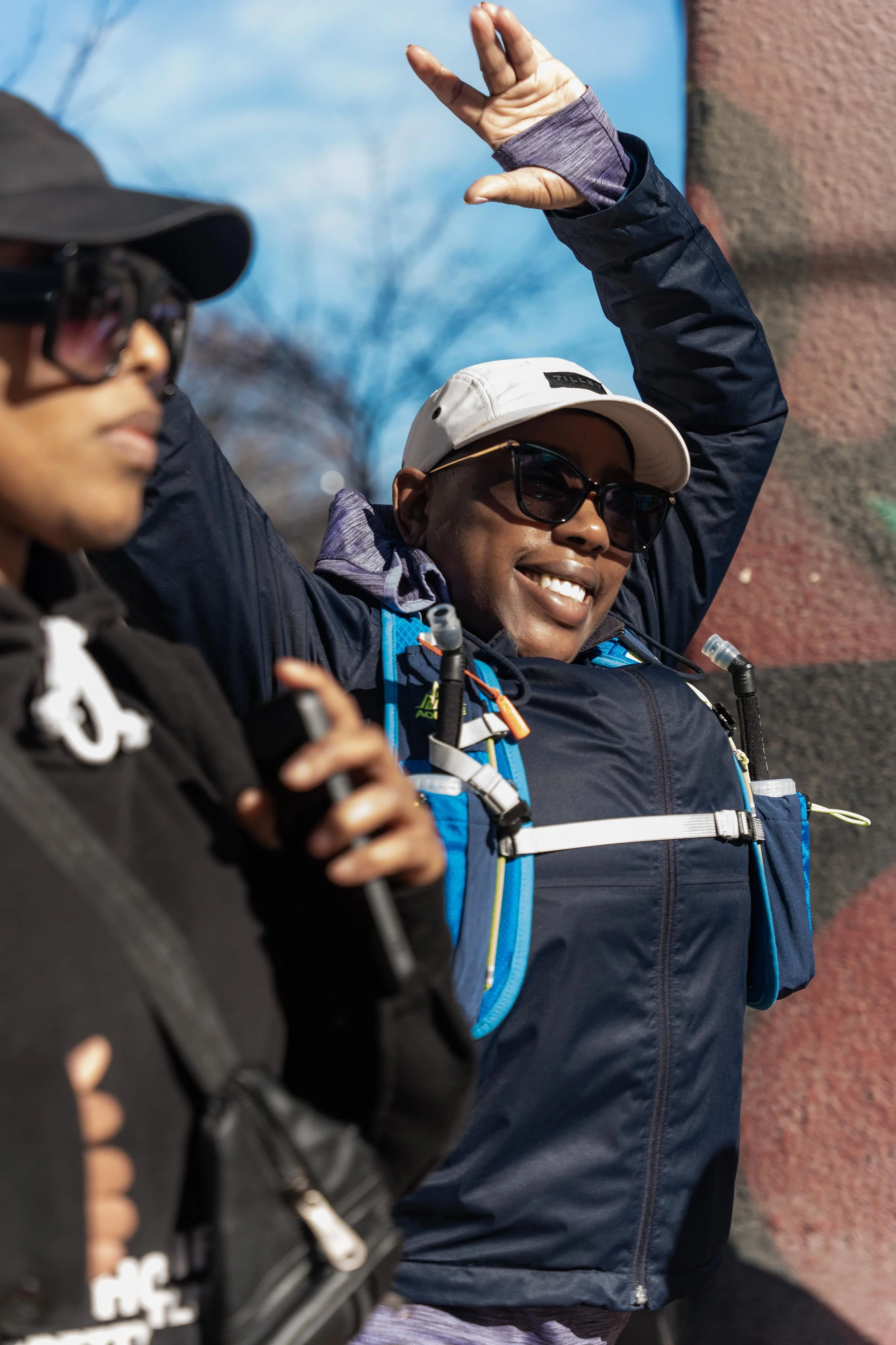 Two people outdoors, one smiling and stretching with an arm raised, wearing sunglasses, a beige cap, and a dark jacket; the other person in the foreground, wearing a black jacket and cap, holding a microphone.