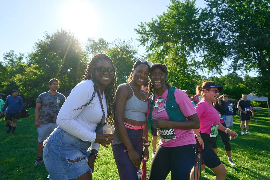 Three women smiling together at a park during a sunny outdoor event, with other people in the background.