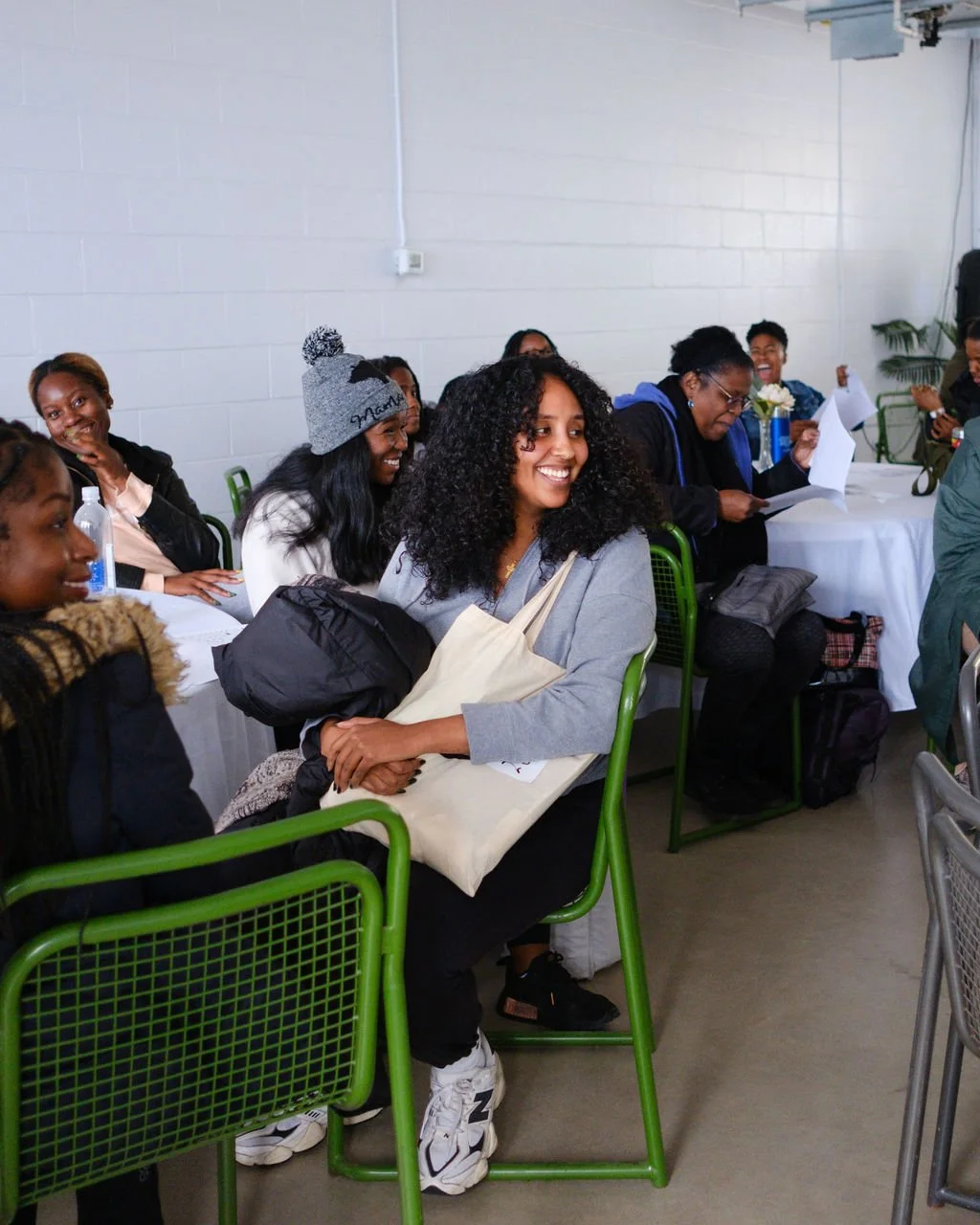 A group of diverse women sitting at a table, smiling and engaging in a gathering or meeting in a bright room with white walls and green chairs.